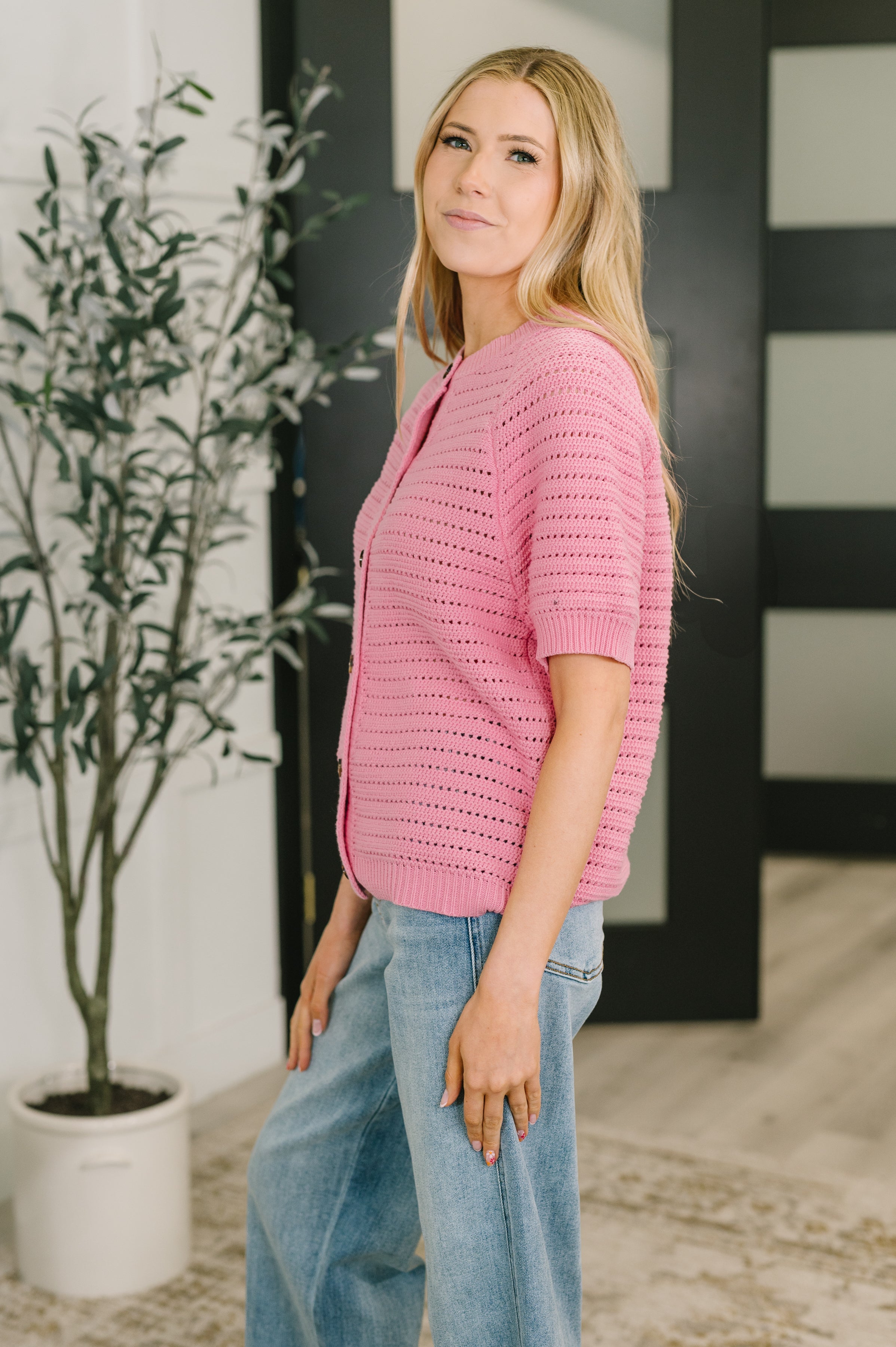 Woman wearing a pink knit button down sweater with short sleeves and light wash wide leg jeans in a bright indoor setting