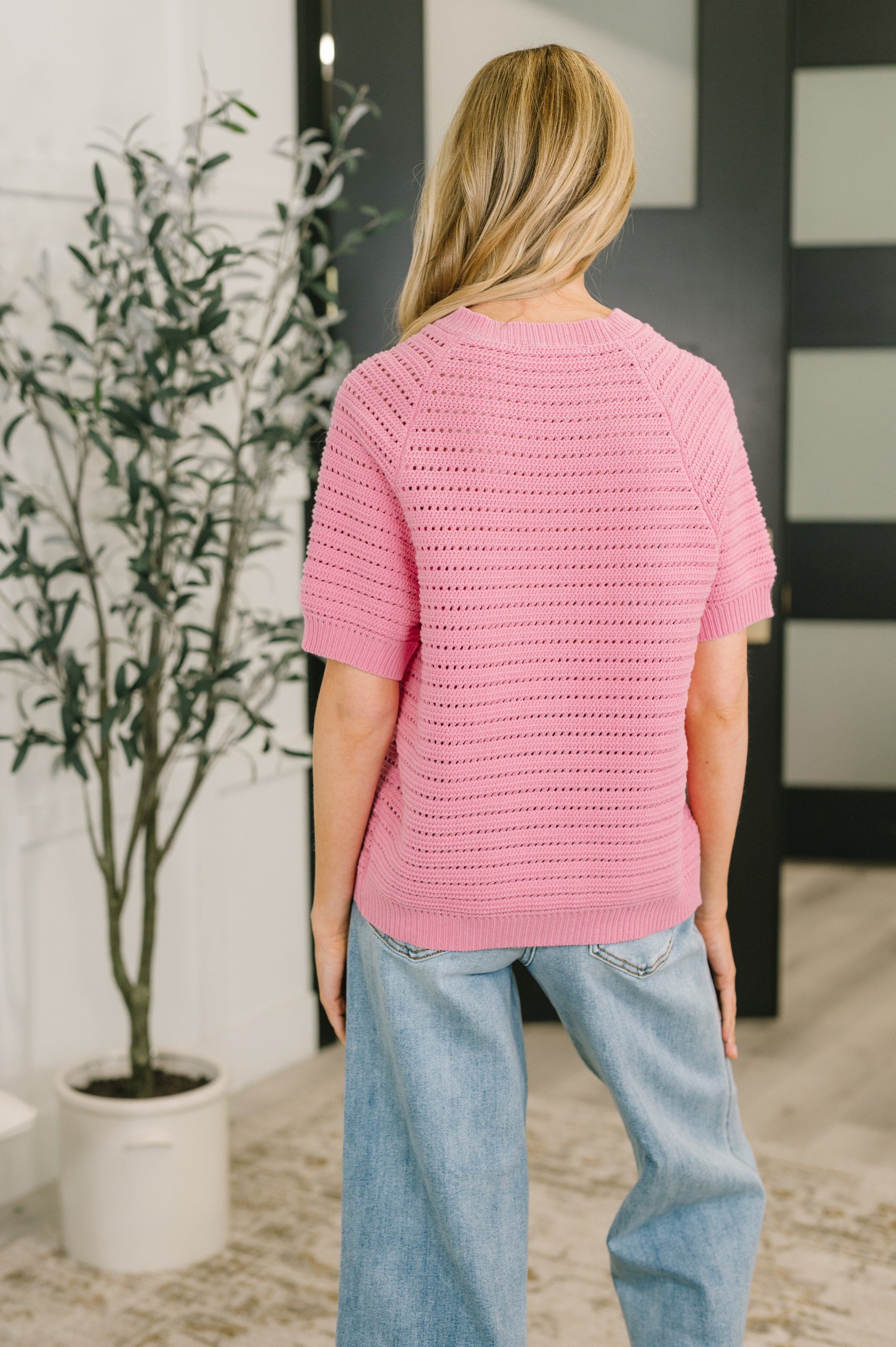 Back view of a woman wearing a pink knit short sleeve button down sweater with a ribbed neckline and light wash jeans indoors