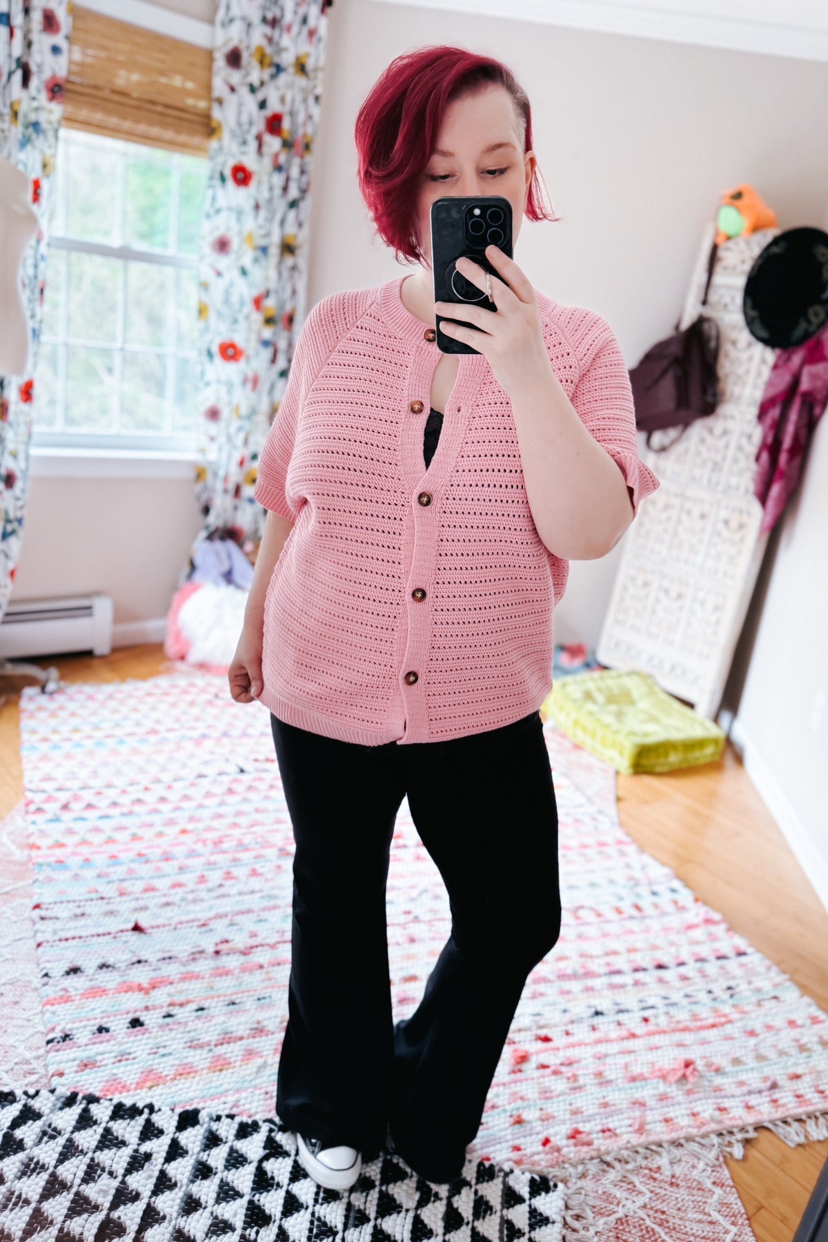 Woman taking a mirror selfie in a pink knit button front sweater with black pants and white sneakers in a colorful bedroom