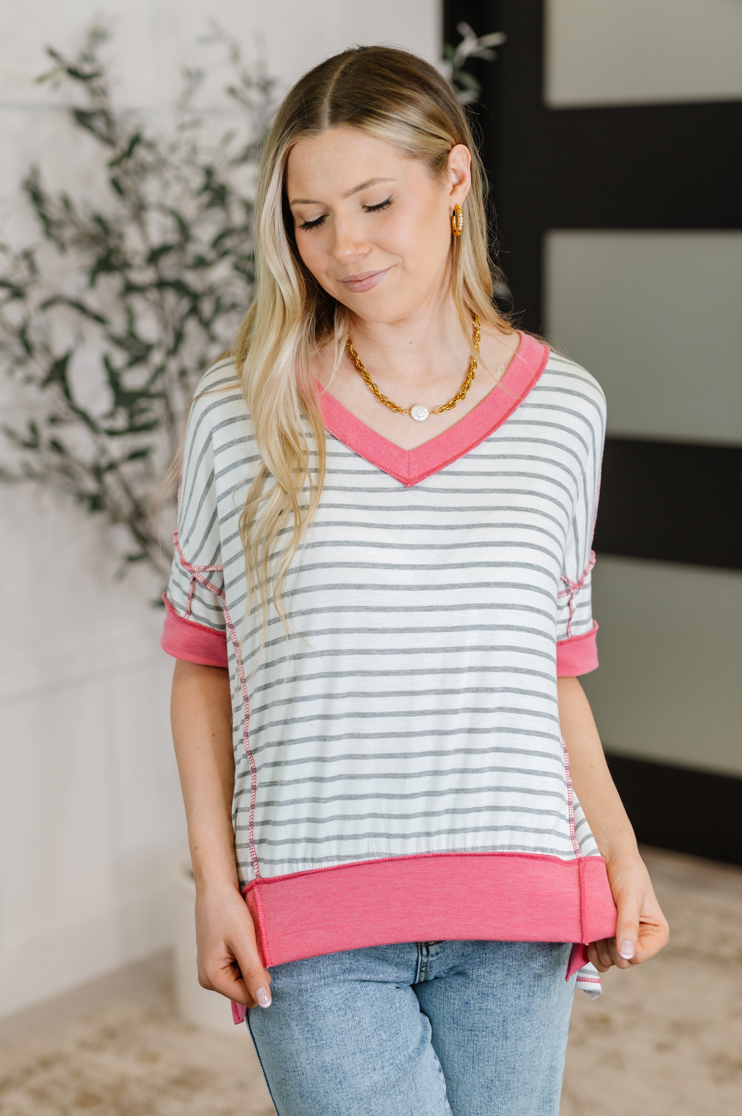 Woman wearing ivory striped V-neck top with bright pink trim and rolled sleeves paired with light blue jeans indoors