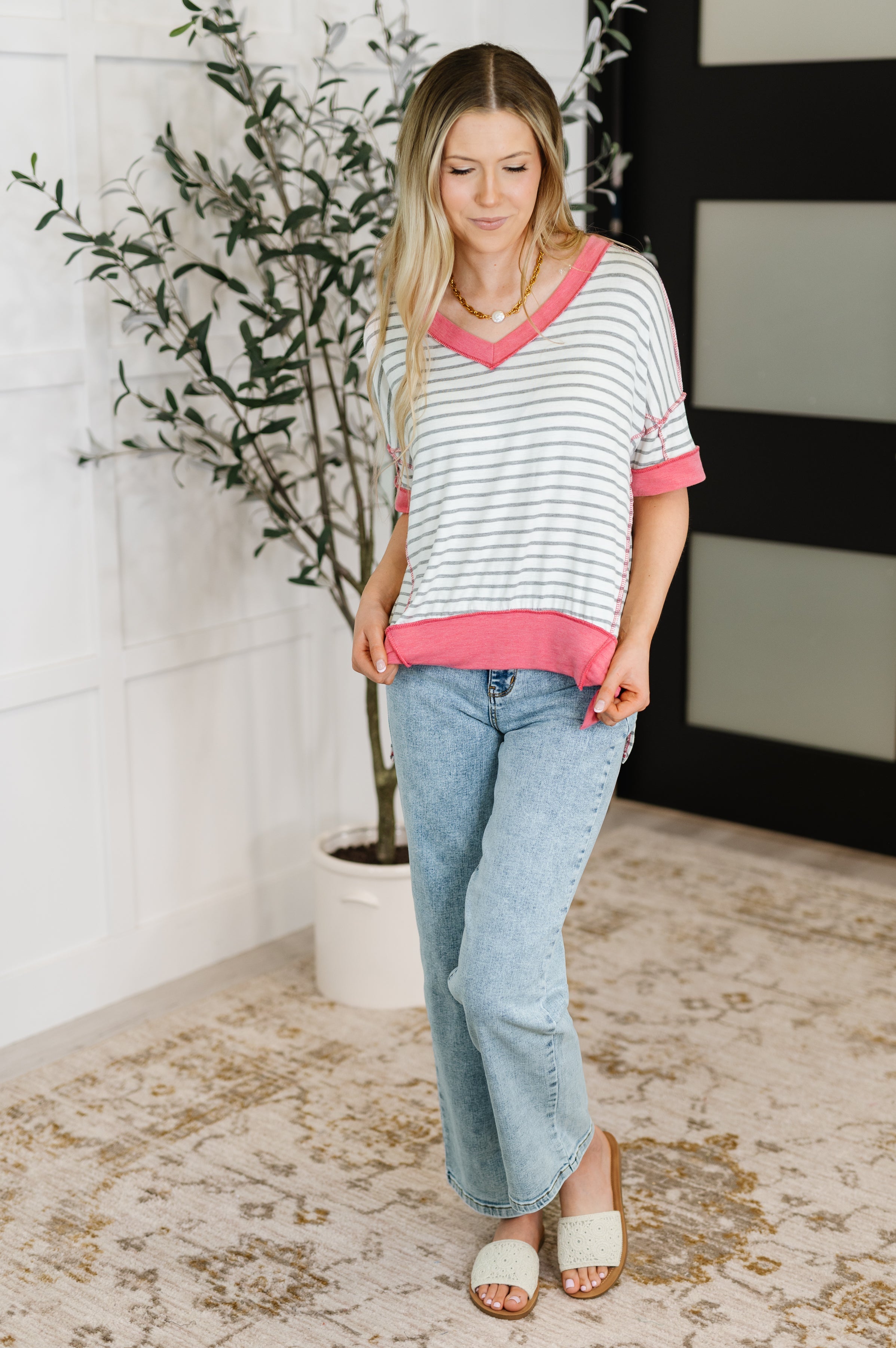 Woman wearing ivory striped V-neck top with coral trim paired with light wash jeans and slide sandals indoors