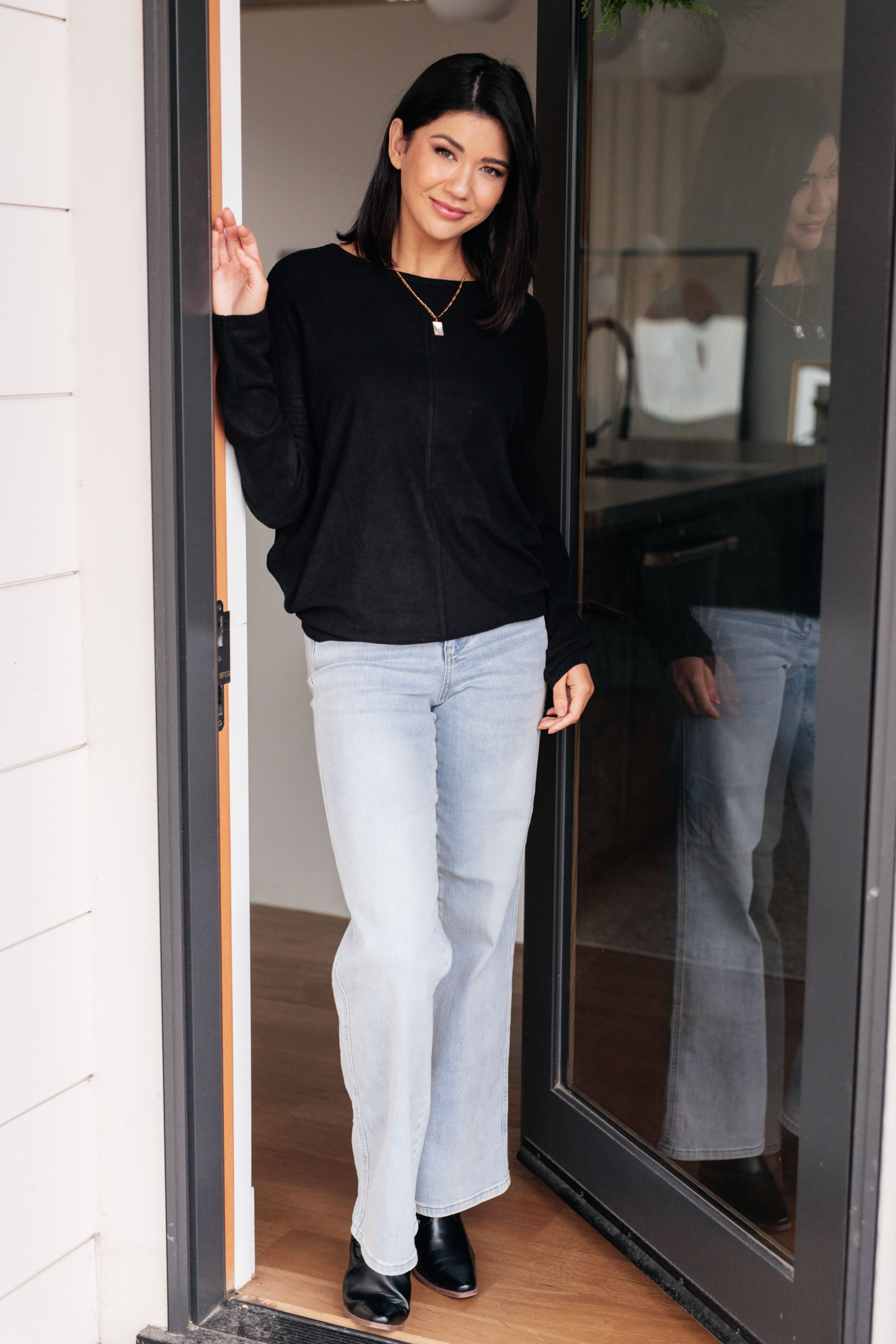 Woman wearing a black dolman long-sleeve top, light blue jeans and black boots standing in a doorway.