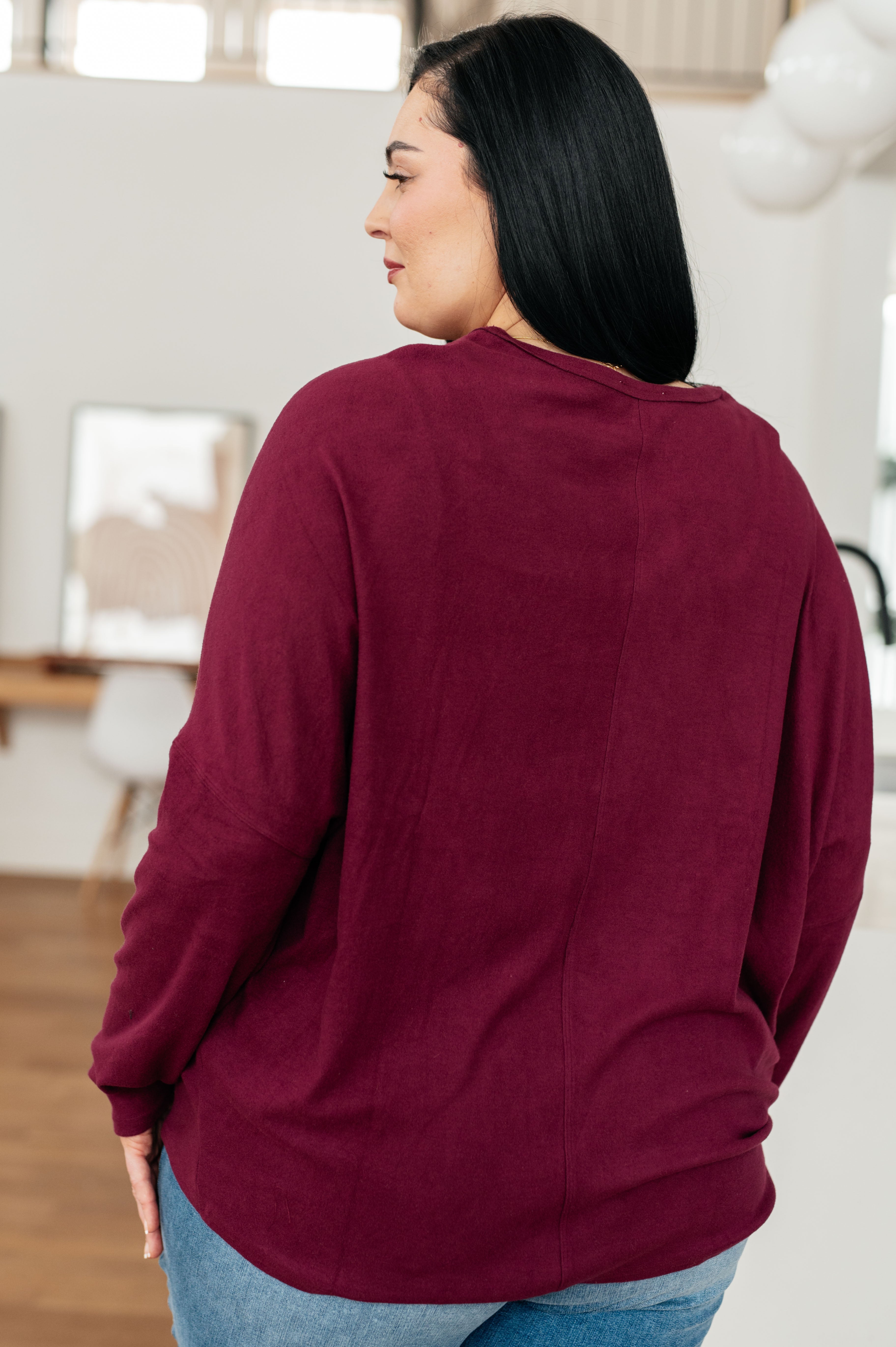 Side profile of woman with long black hair wearing wine-colored dolman sleeve top and blue jeans indoors