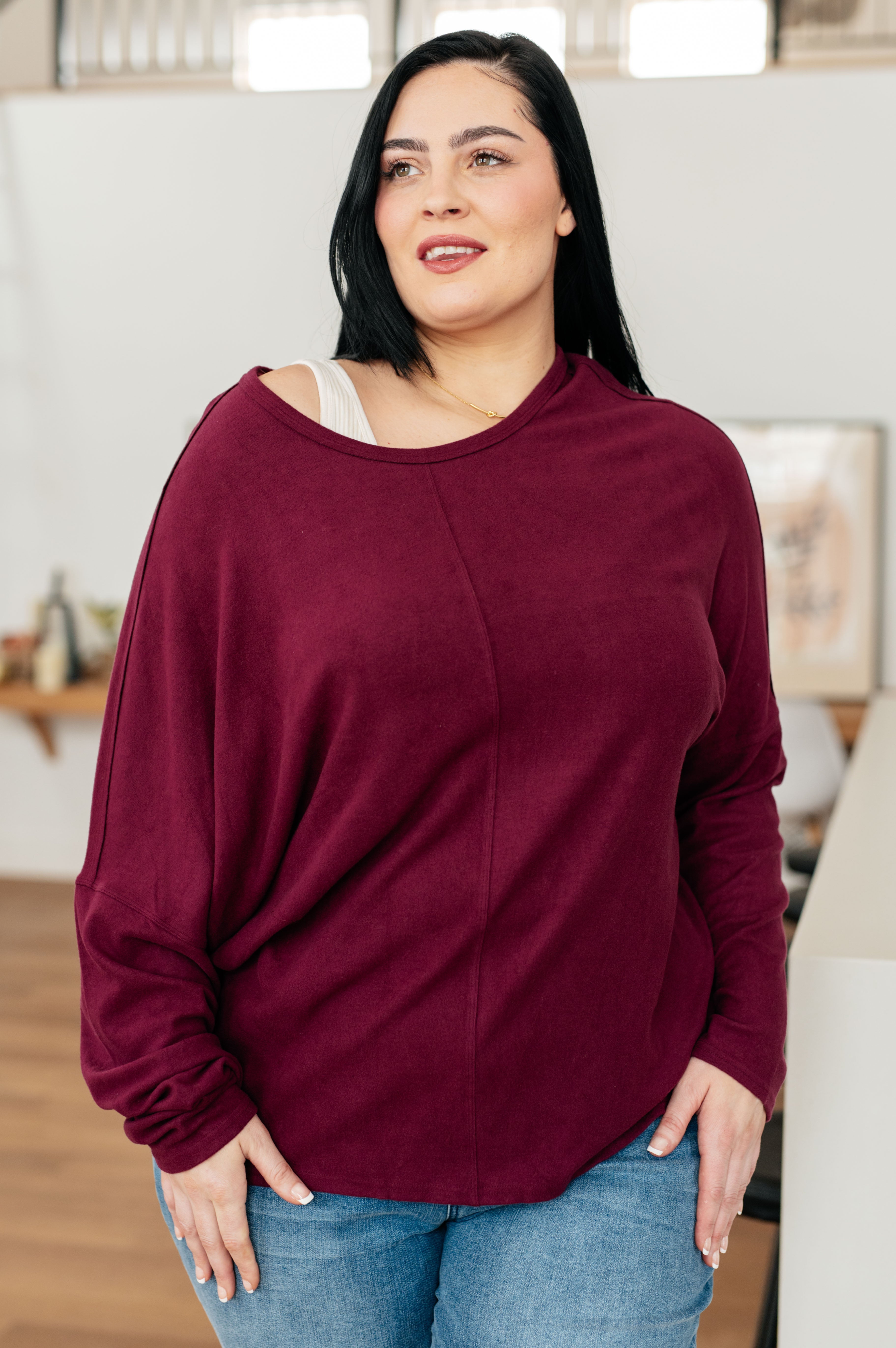 Plus size woman with long black hair wearing loose wine-colored dolman sleeve top and light blue jeans indoors