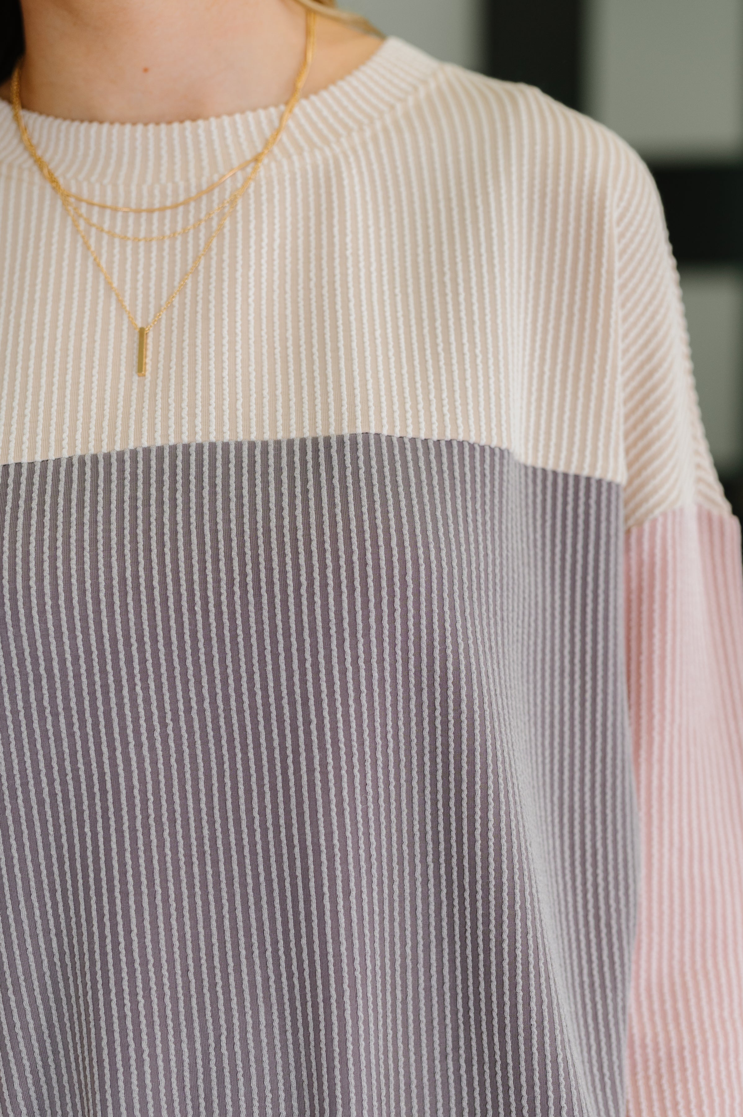 Close-up of ribbed knit sweater in cream, gray, and pink tones worn with layered gold necklaces indoors