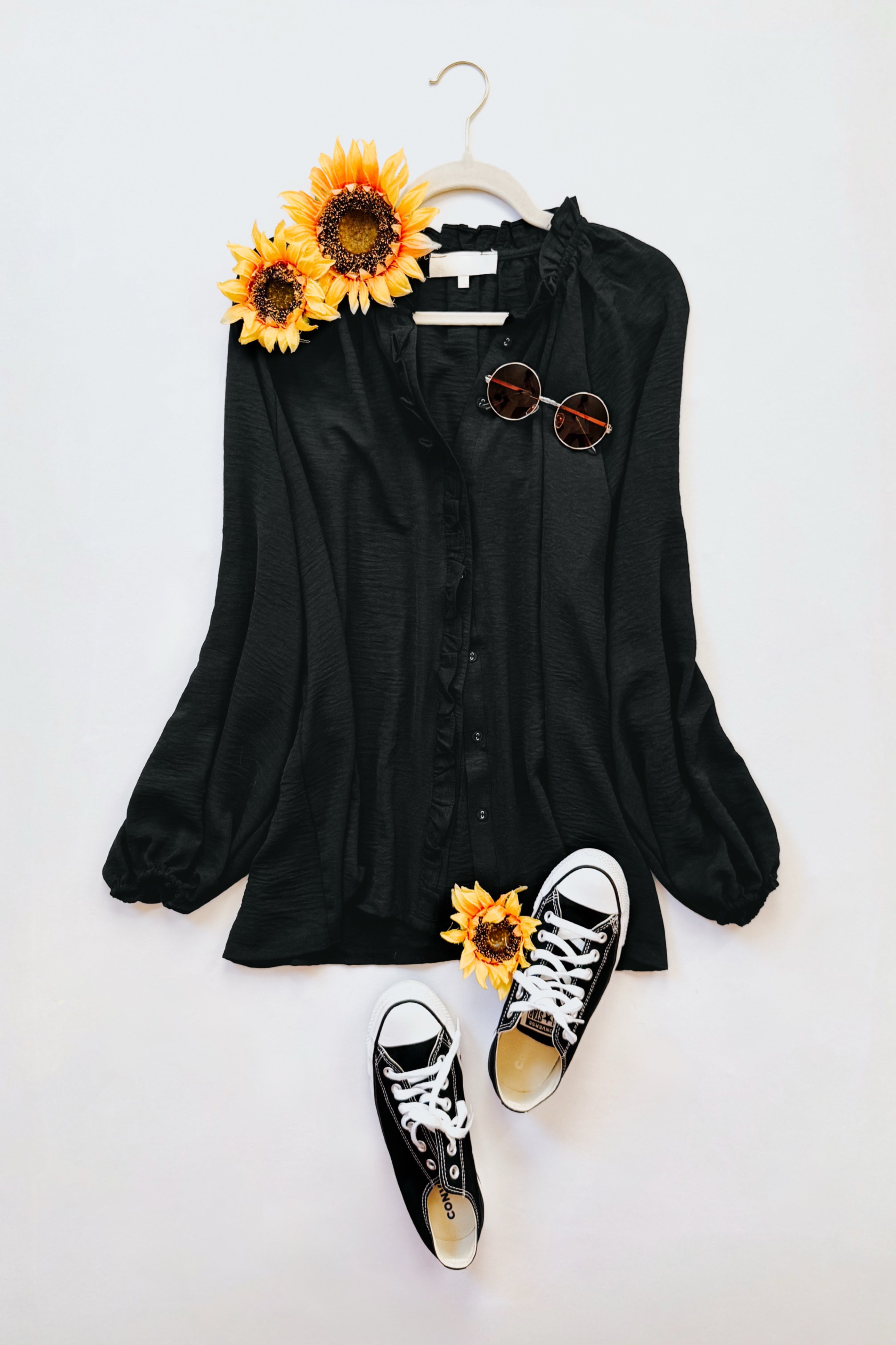 Flay featuring a black button up ruffled blouse on a hanger with sunflowers and black sneakers on a white background