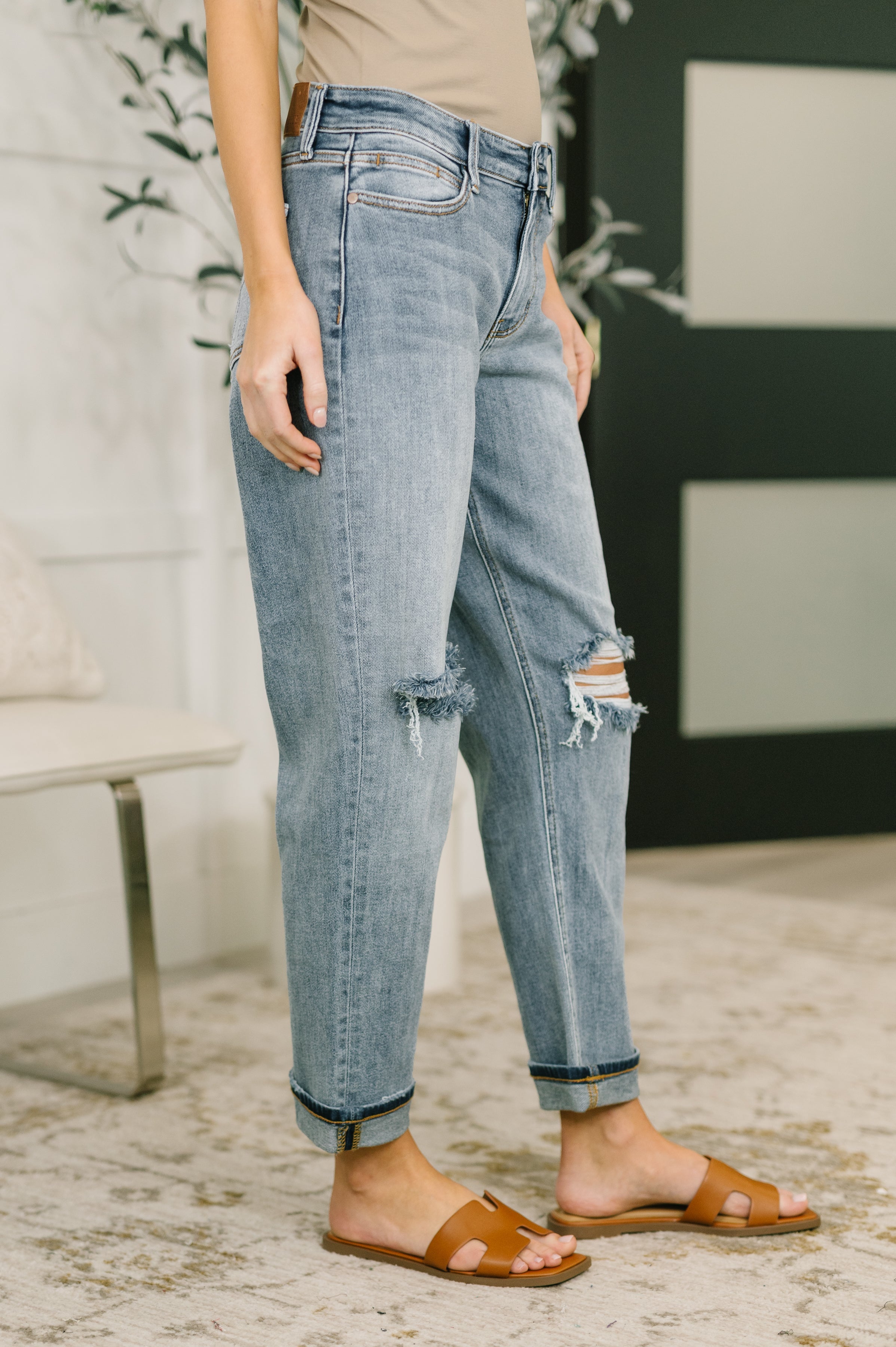 Woman modeling blue distressed denim jeans with rolled cuffs and brown slide sandals indoors