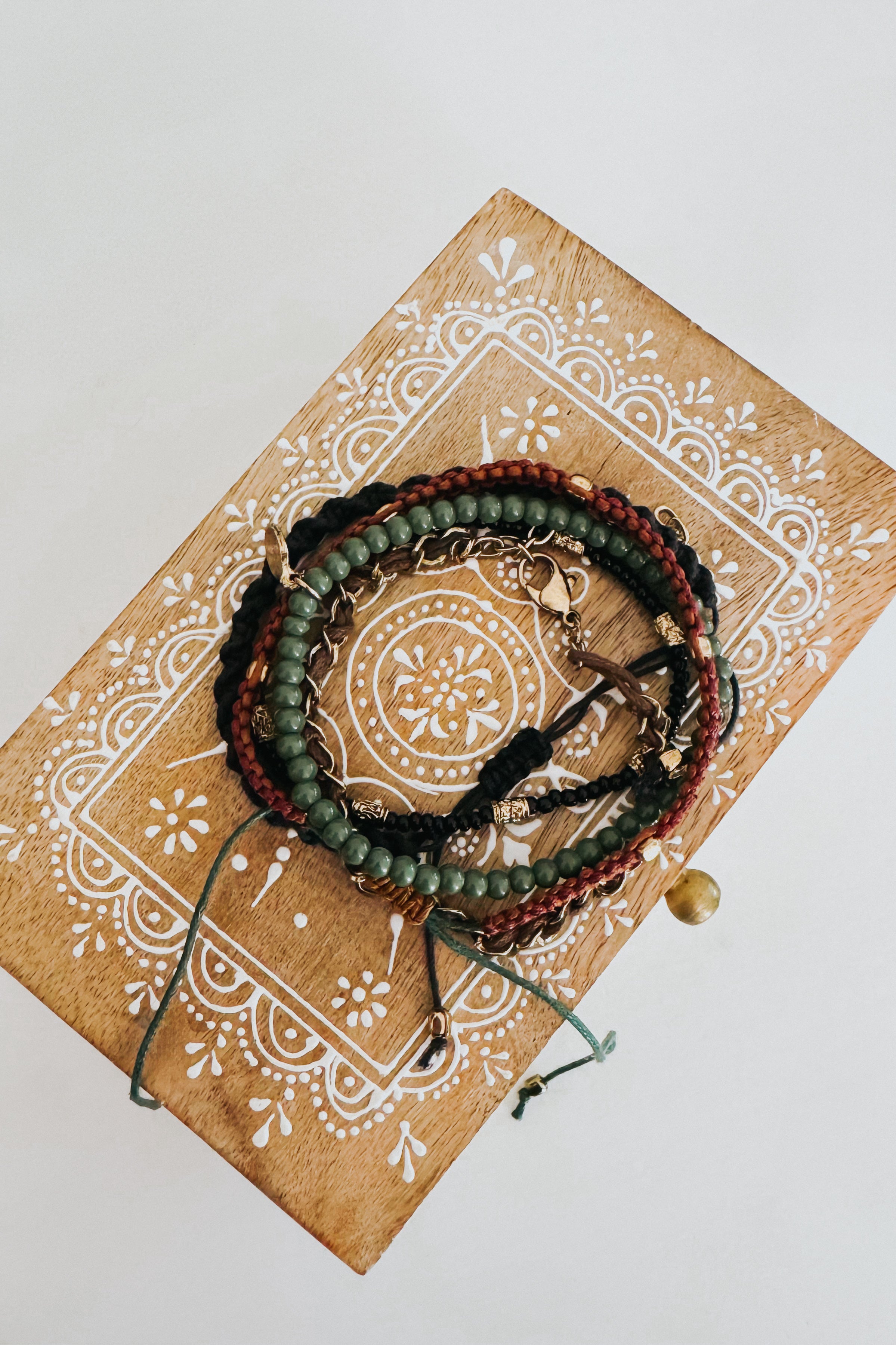 Coiled Ember Stackable bracelet set with green beads, braided cords in black and brown, and gold clasp on decorative wooden box