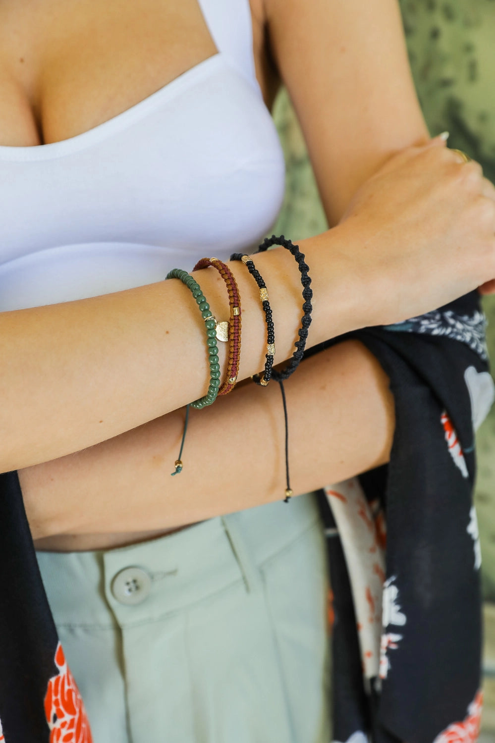 Close-up of a wrist wearing four Ember Stackable bracelets with green beads, brown and black braided cords, and gold accents