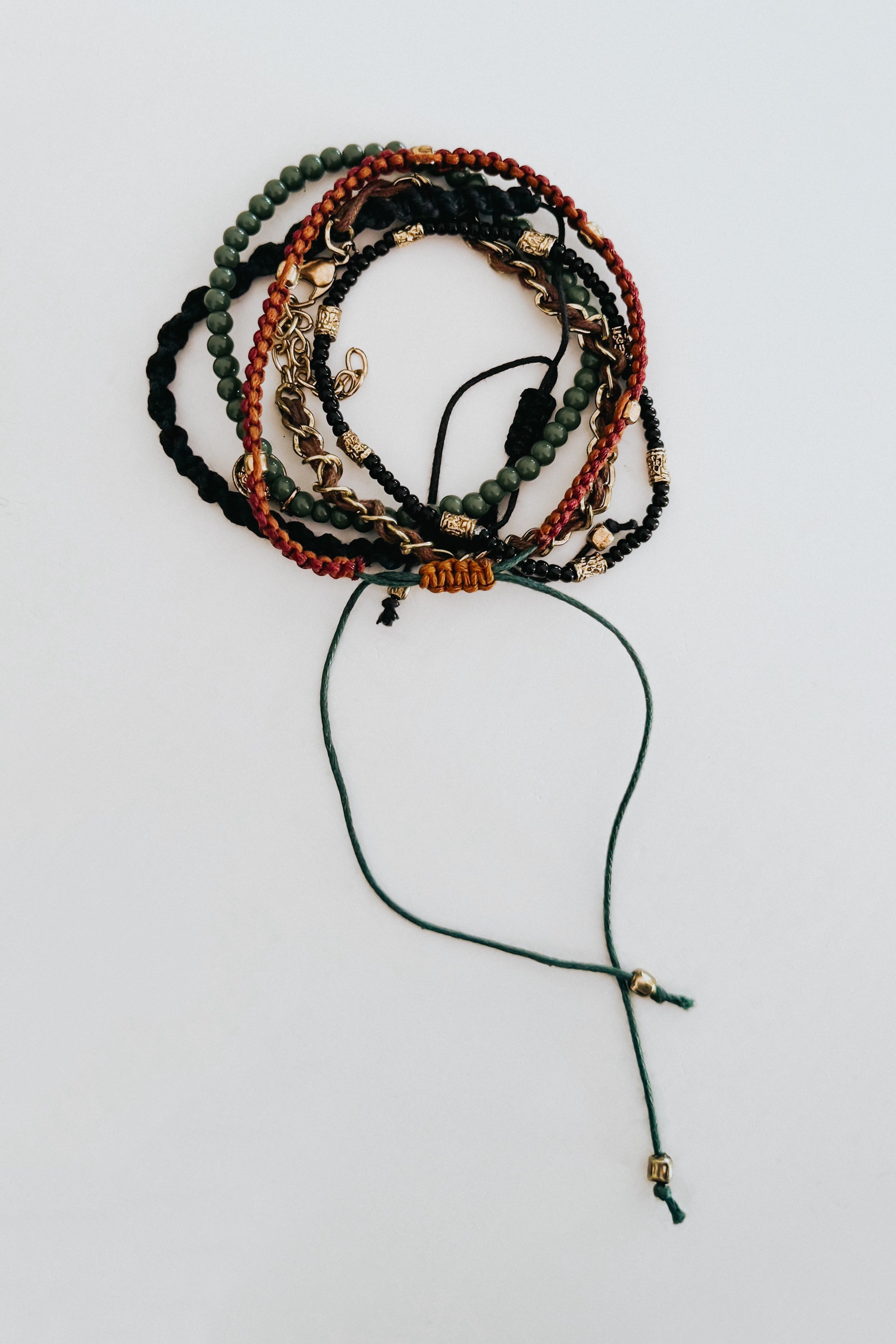 Set of stackable Ember bracelets featuring braided cords, green beads, and gold accents on a light background