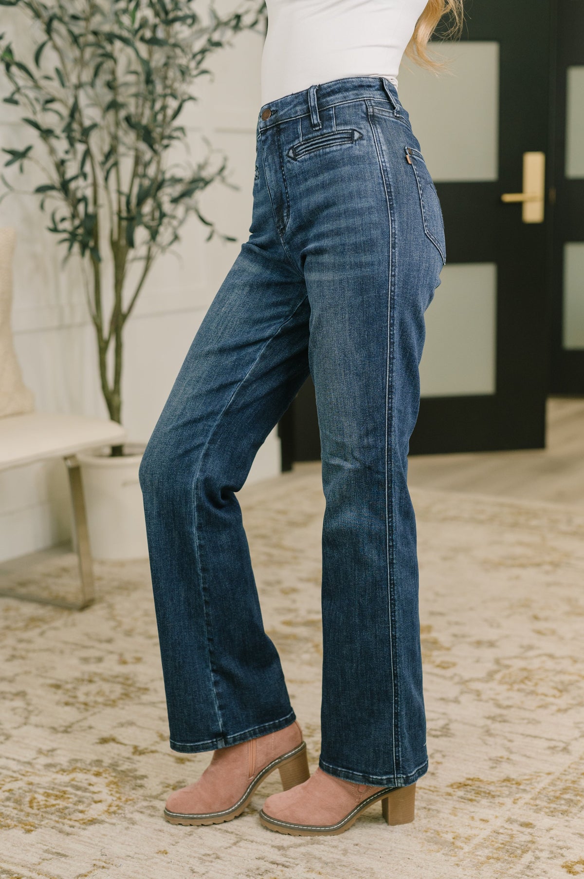 Woman models dark blue high waist bootcut jeans with welt pockets and tan heeled shoes in a stylish modern entryway