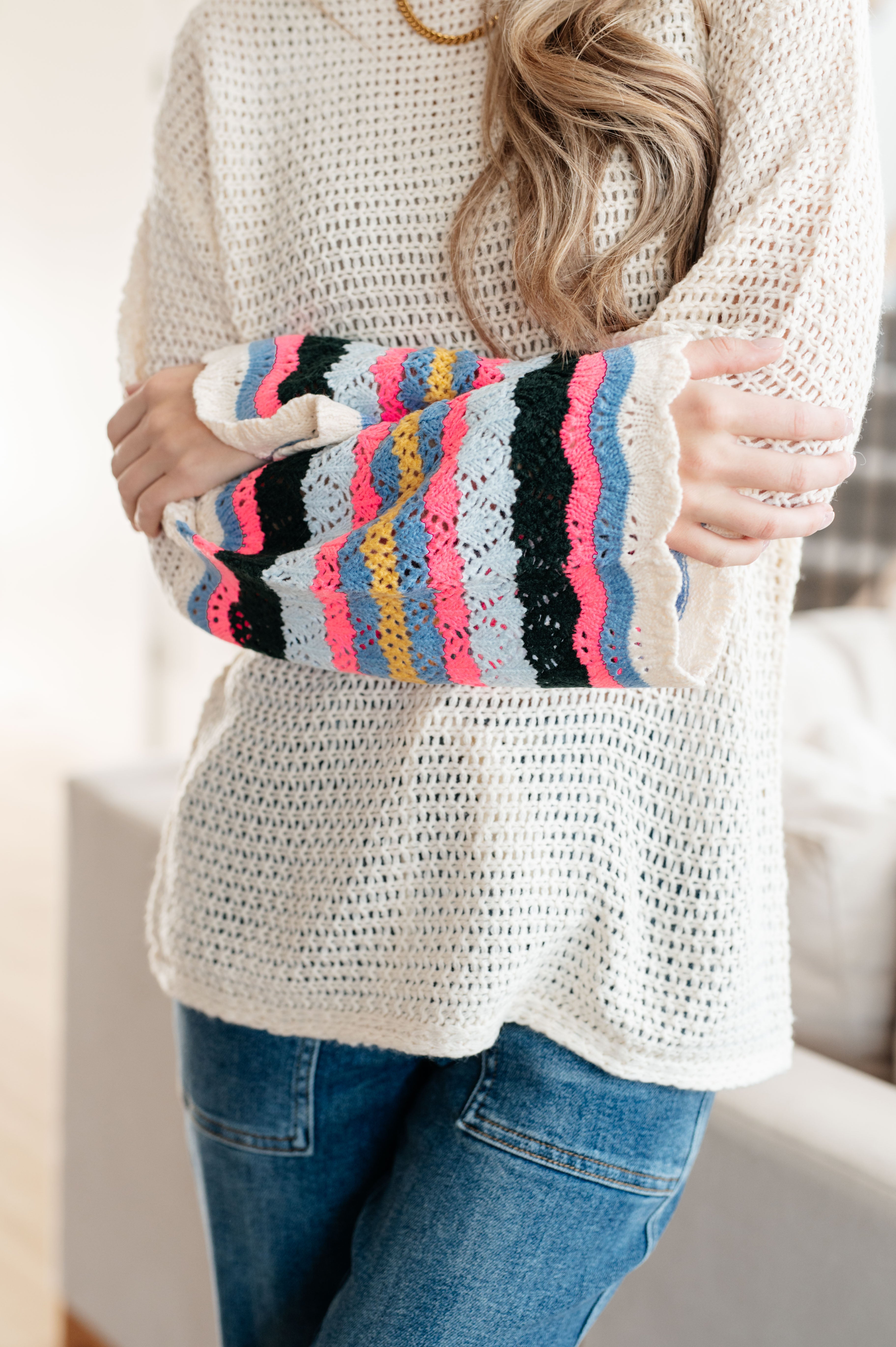 Close-up of woman wearing cream knit sweater with colorful striped lace sleeves and blue jeans indoors
