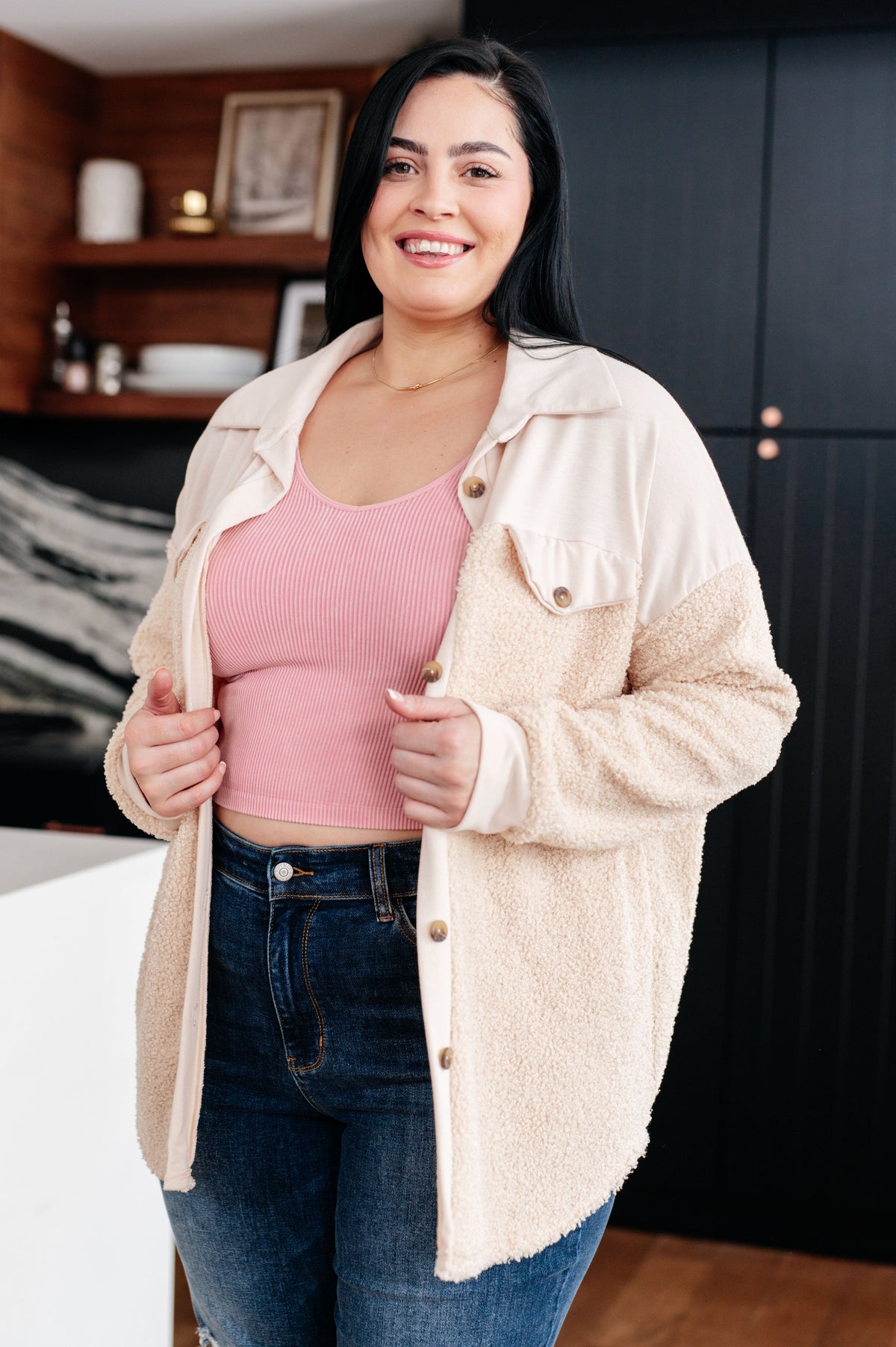 Smiling woman wearing a cream sherpa shacket with faux chest pockets over a pink ribbed tank and dark jeans indoors