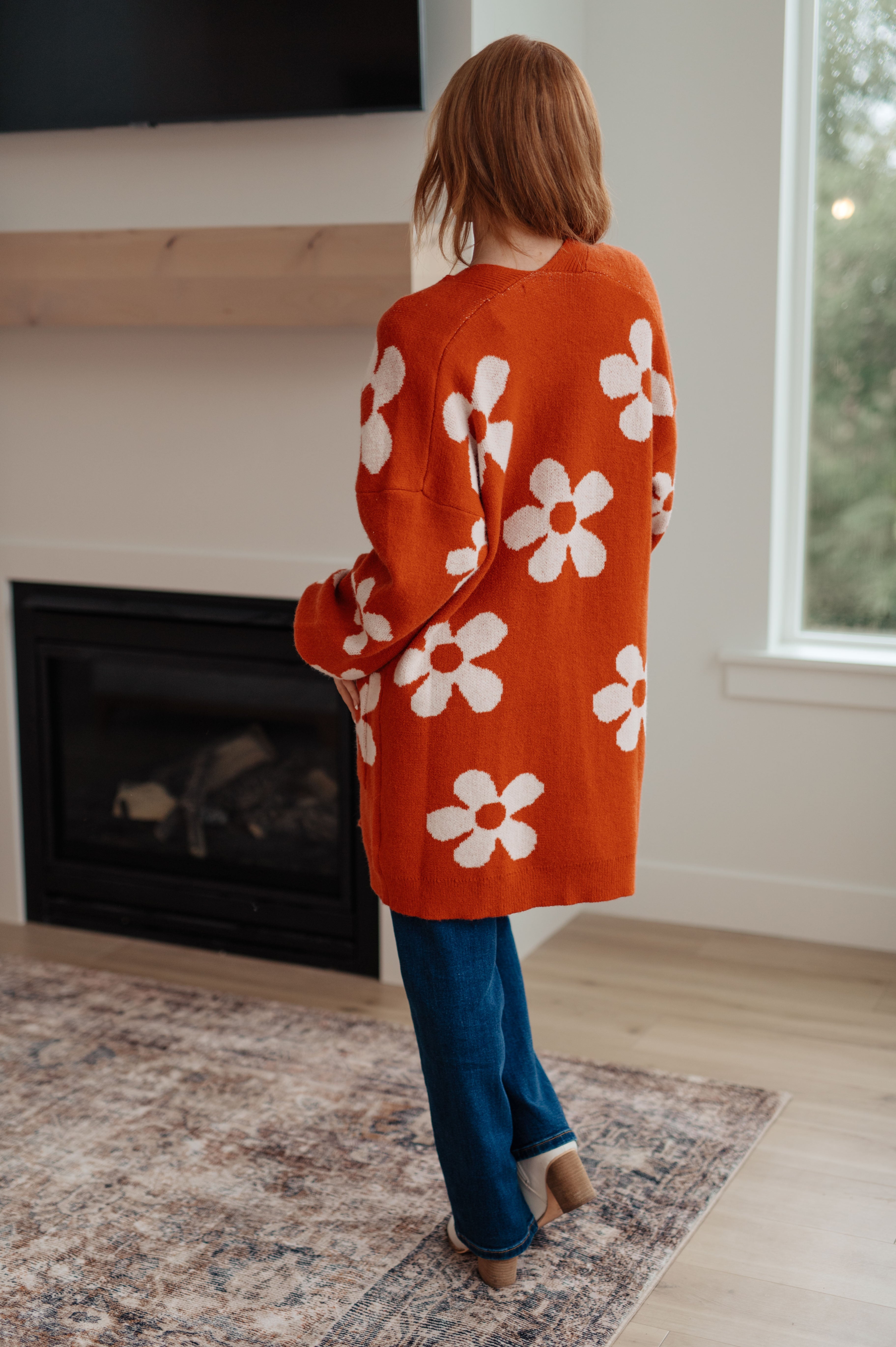 Woman standing indoors wearing burnt orange cardigan with large white floral print and blue jeans by fireplace