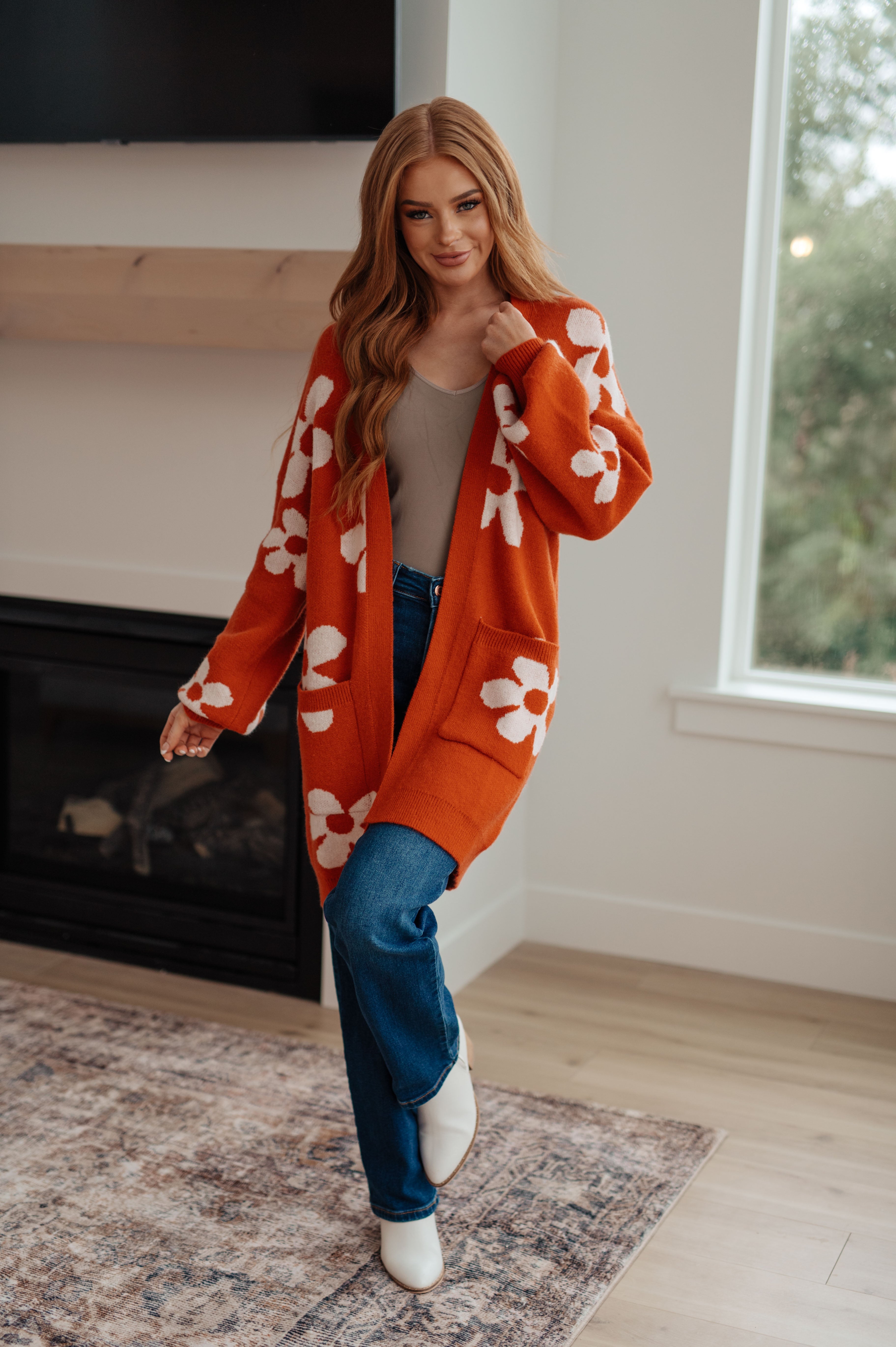 Woman indoors wearing burnt orange floral cardigan over beige top and blue jeans with white ankle boots standing near fireplace