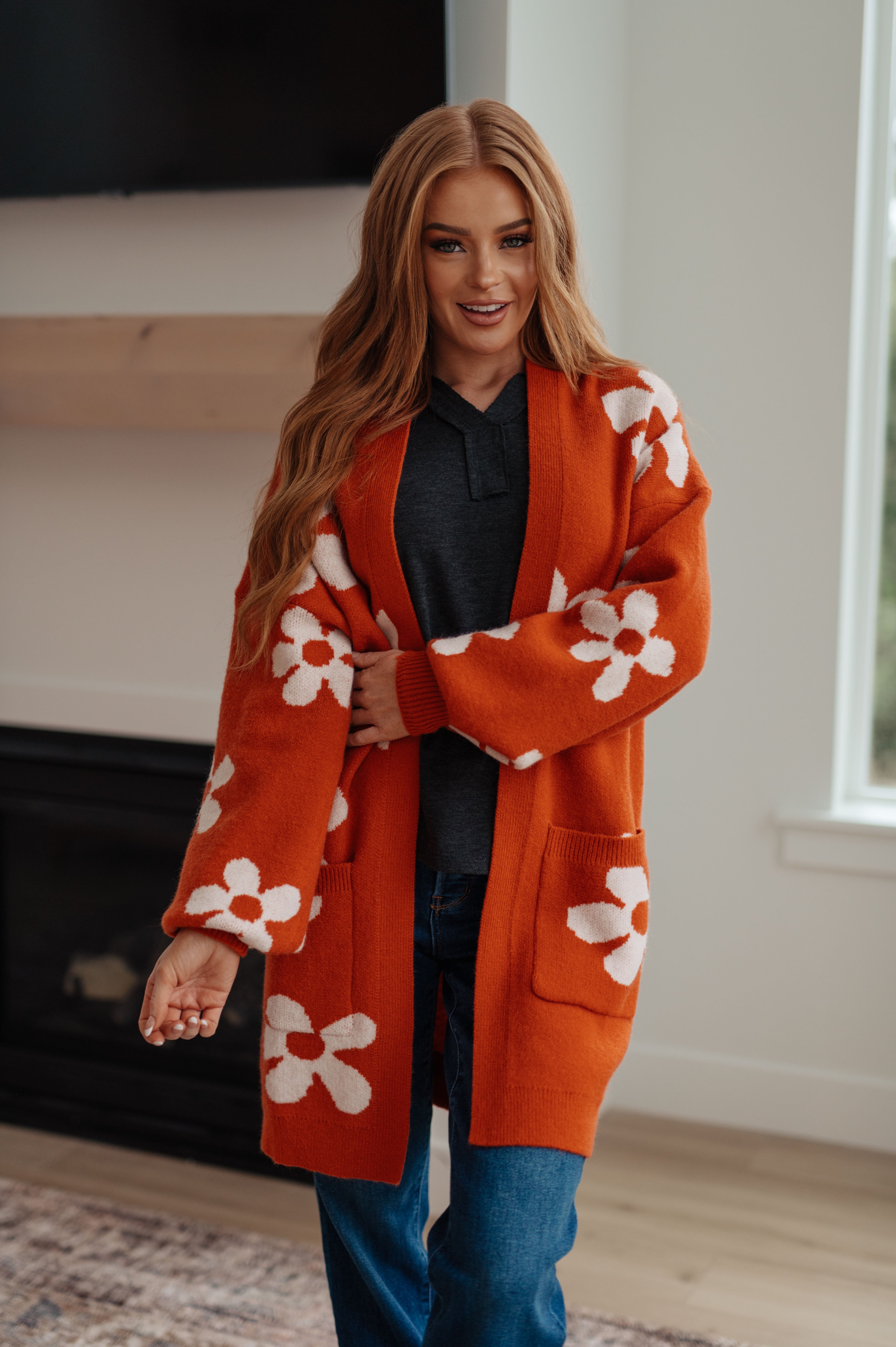 Woman wearing burnt orange floral cardigan with front pockets and dark shirt standing indoors near fireplace