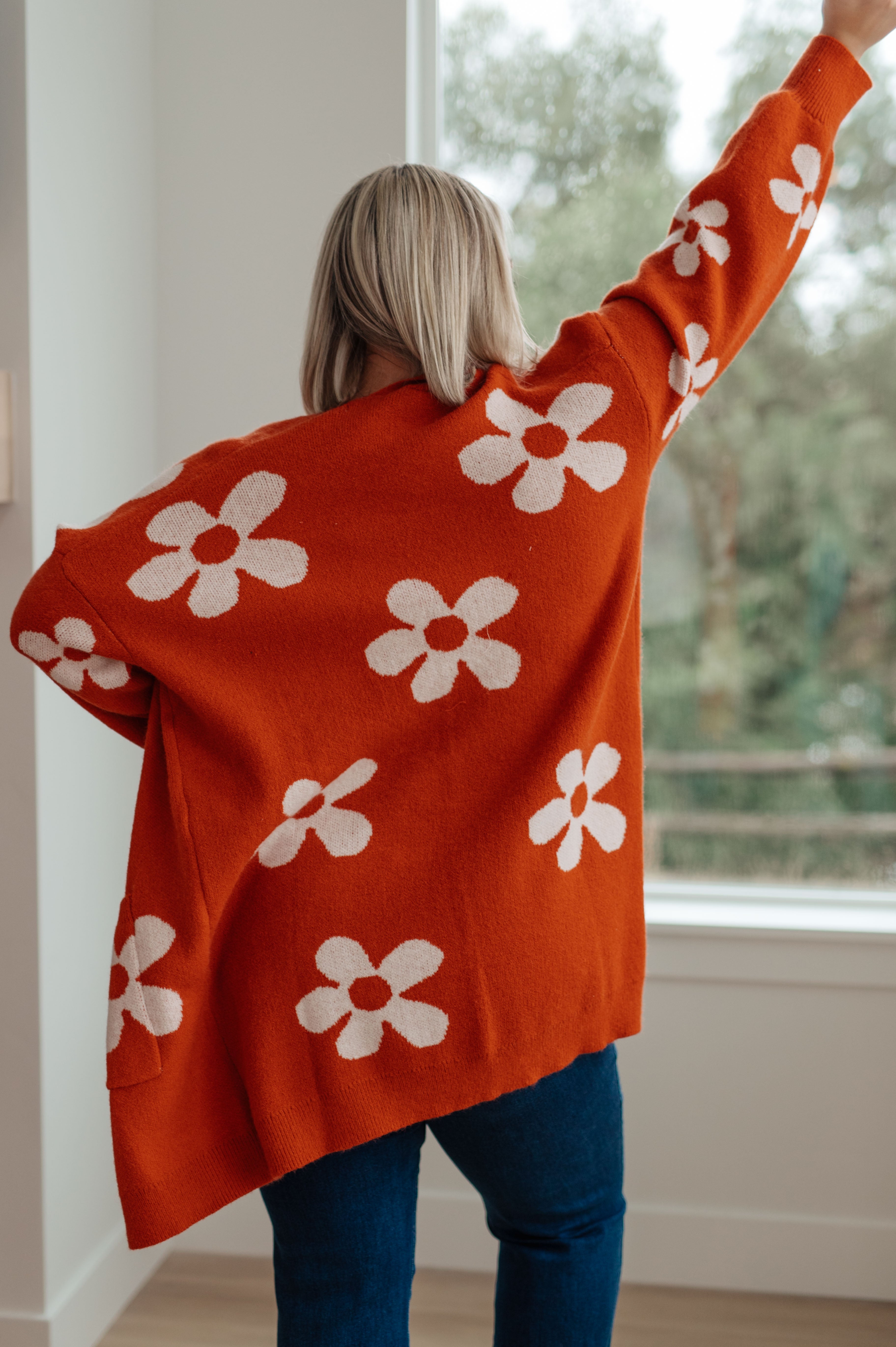 Woman wearing burnt orange cardigan with large white floral pattern standing indoors facing window