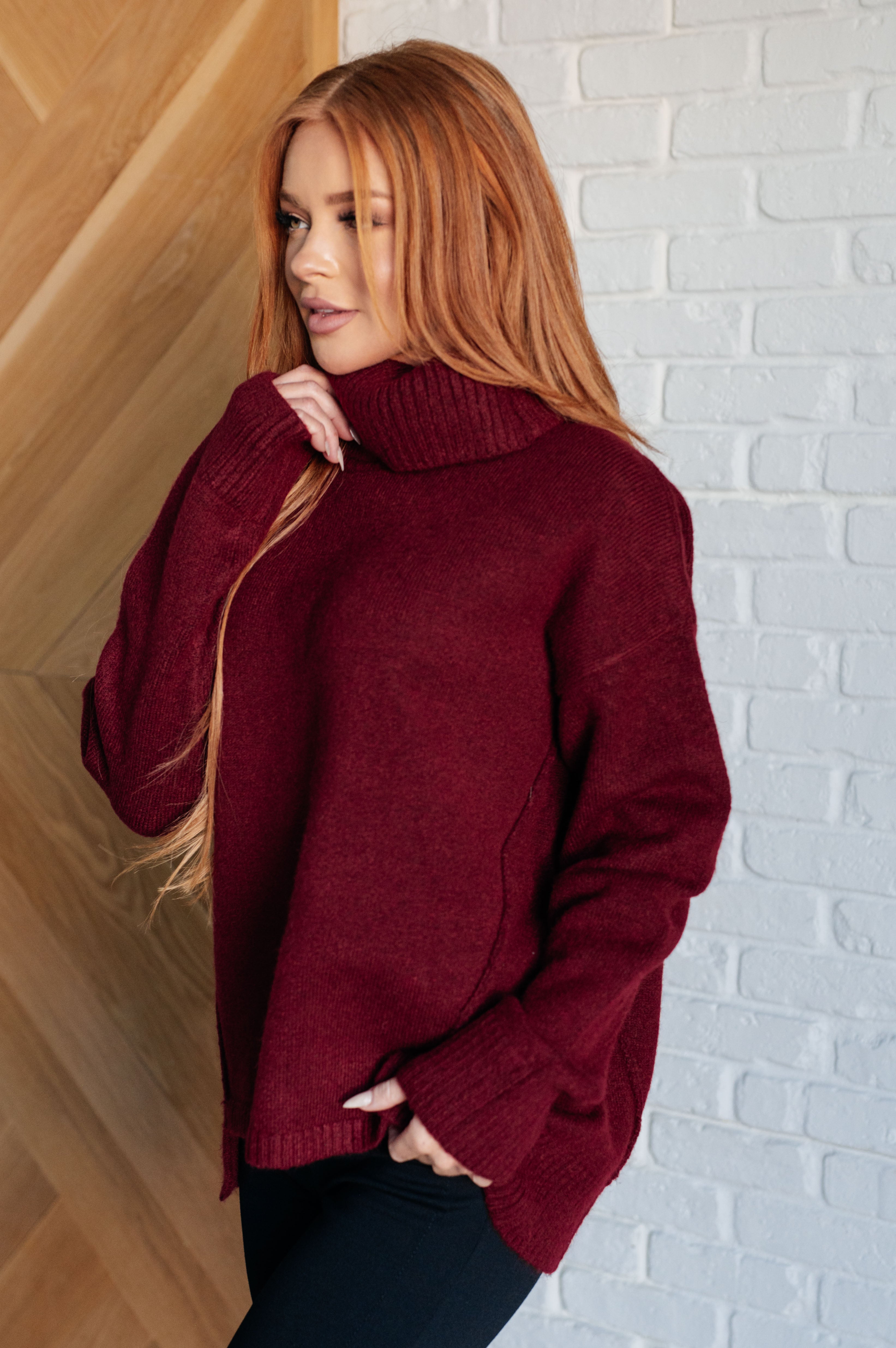 Red-haired woman wearing oversized dark red turtleneck sweater standing indoors near wooden and brick walls