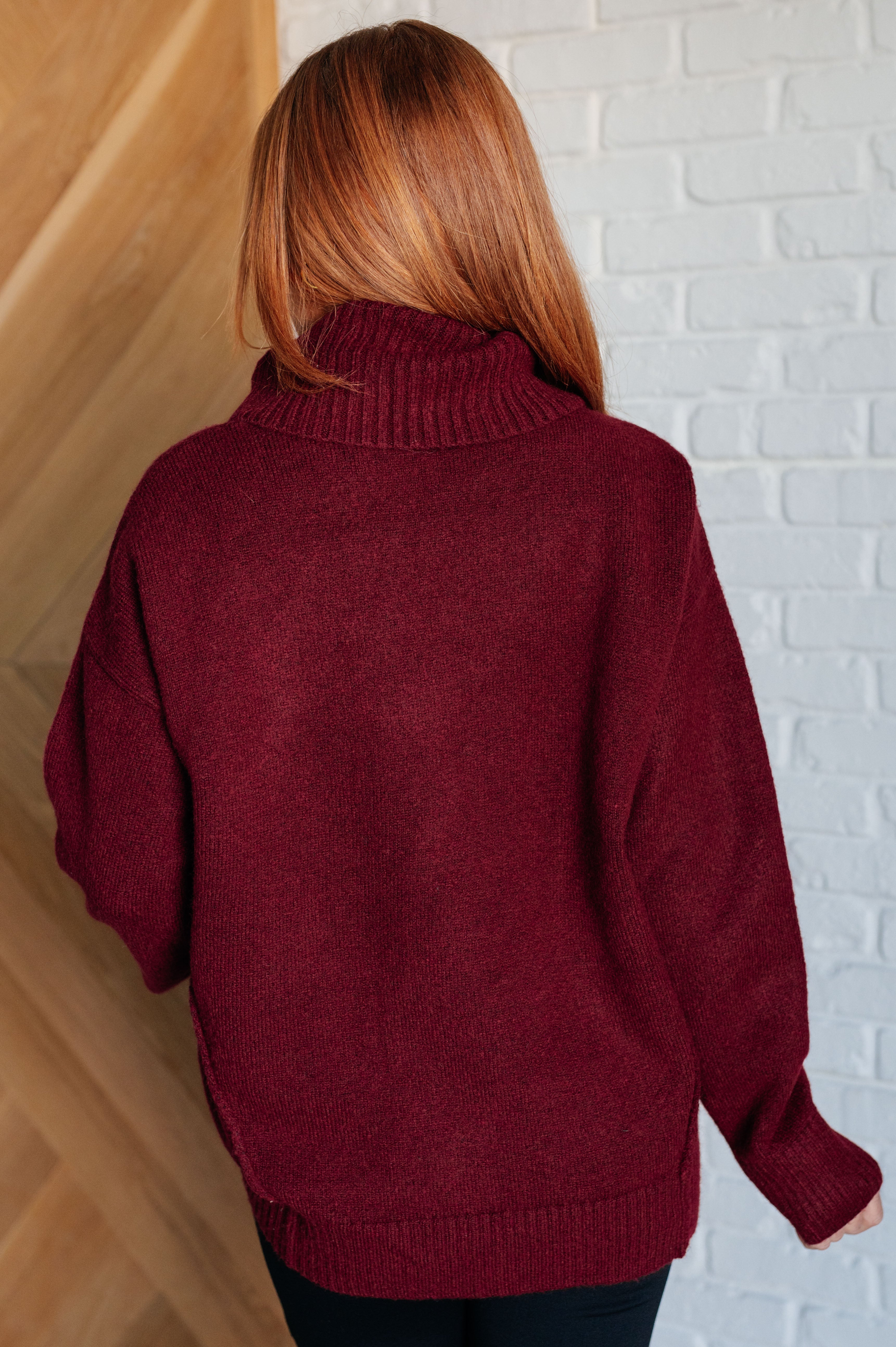 Woman with red hair wearing burgundy turtleneck sweater seen from behind indoors against white brick wall