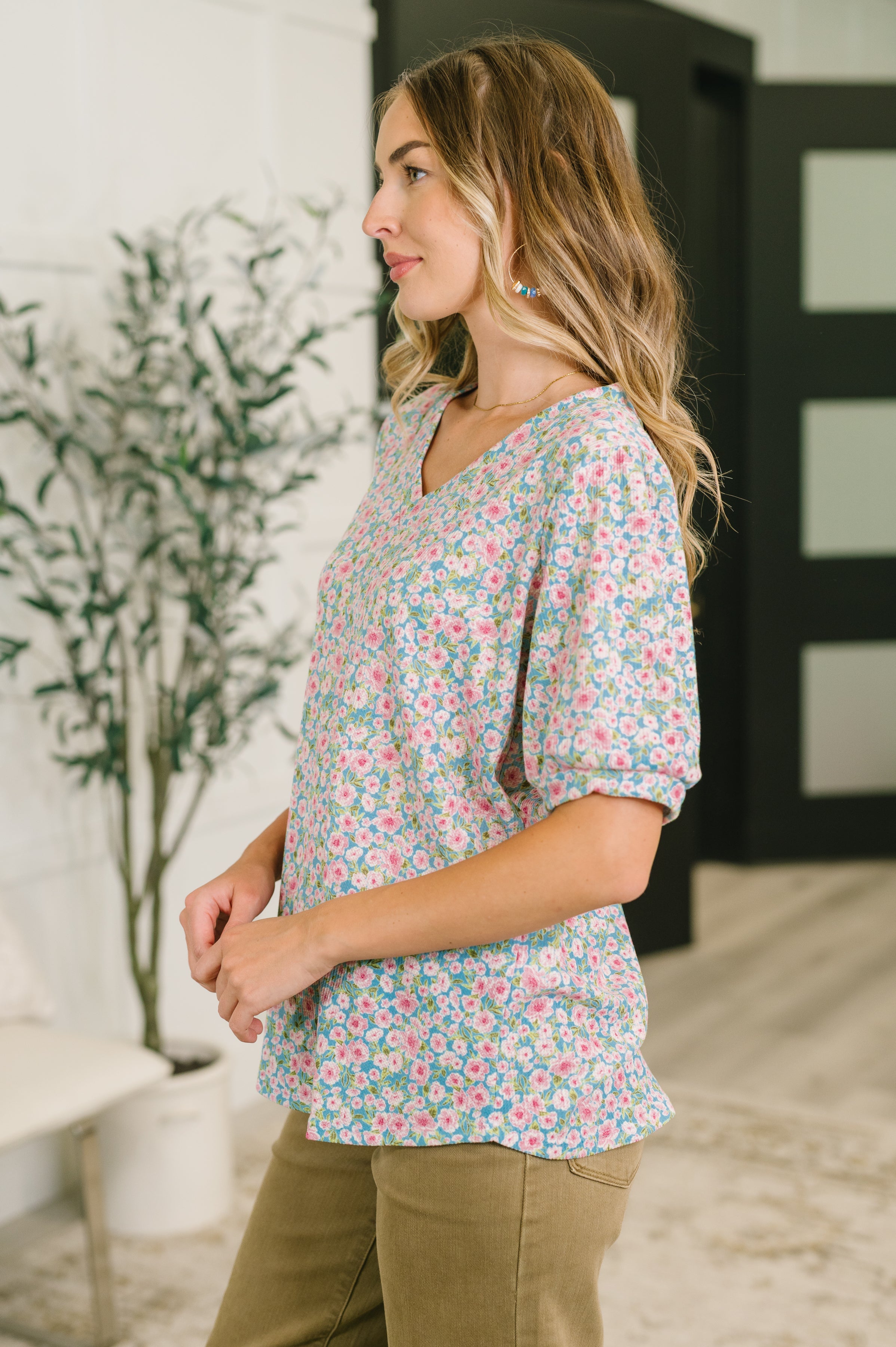 Side profile of woman wearing blue floral puff sleeve blouse with banded V-neckline and khaki pants indoors