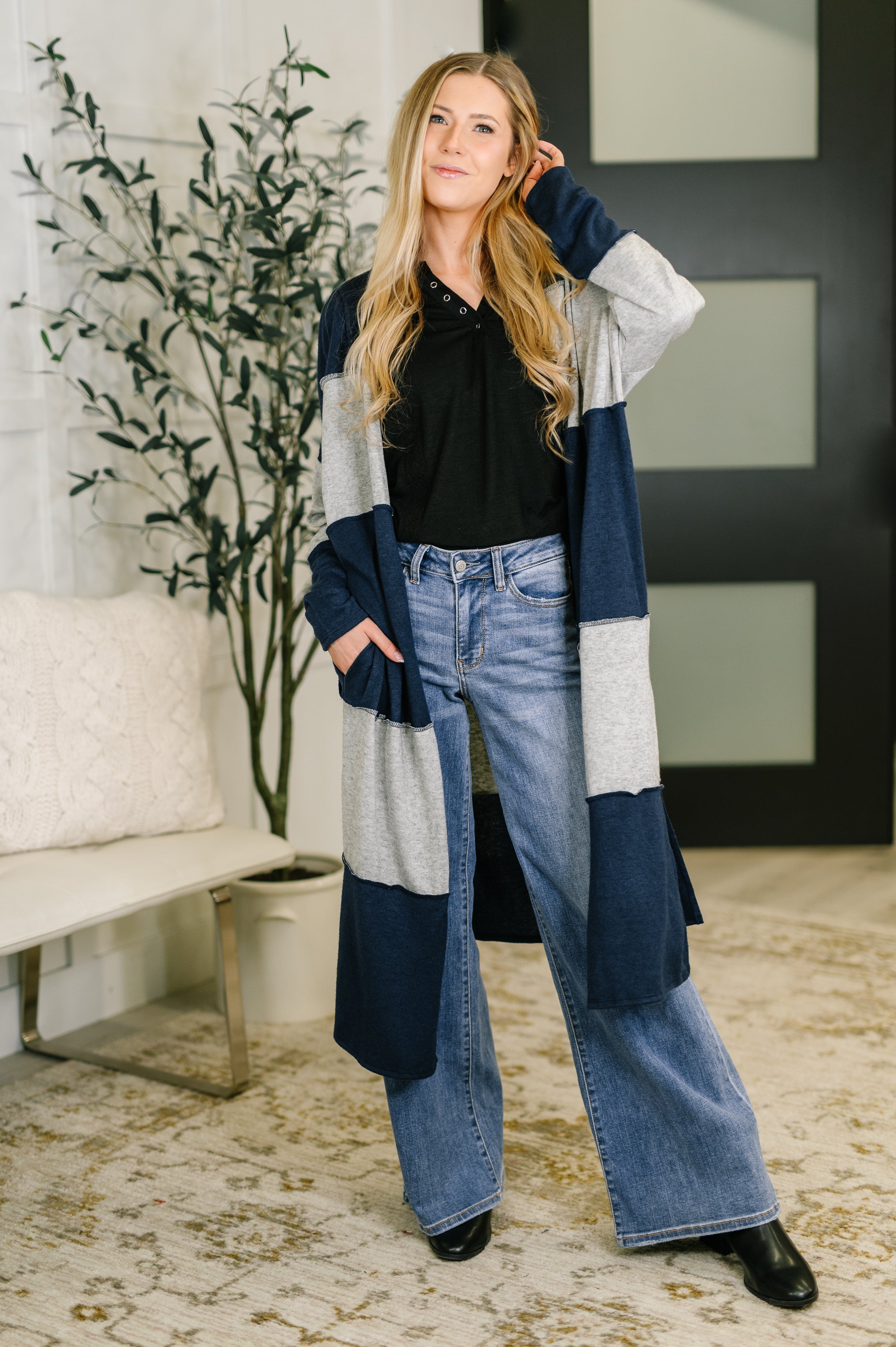 Woman wearing longline open front cardigan with navy and gray color blocking over black top and wide-leg jeans indoors