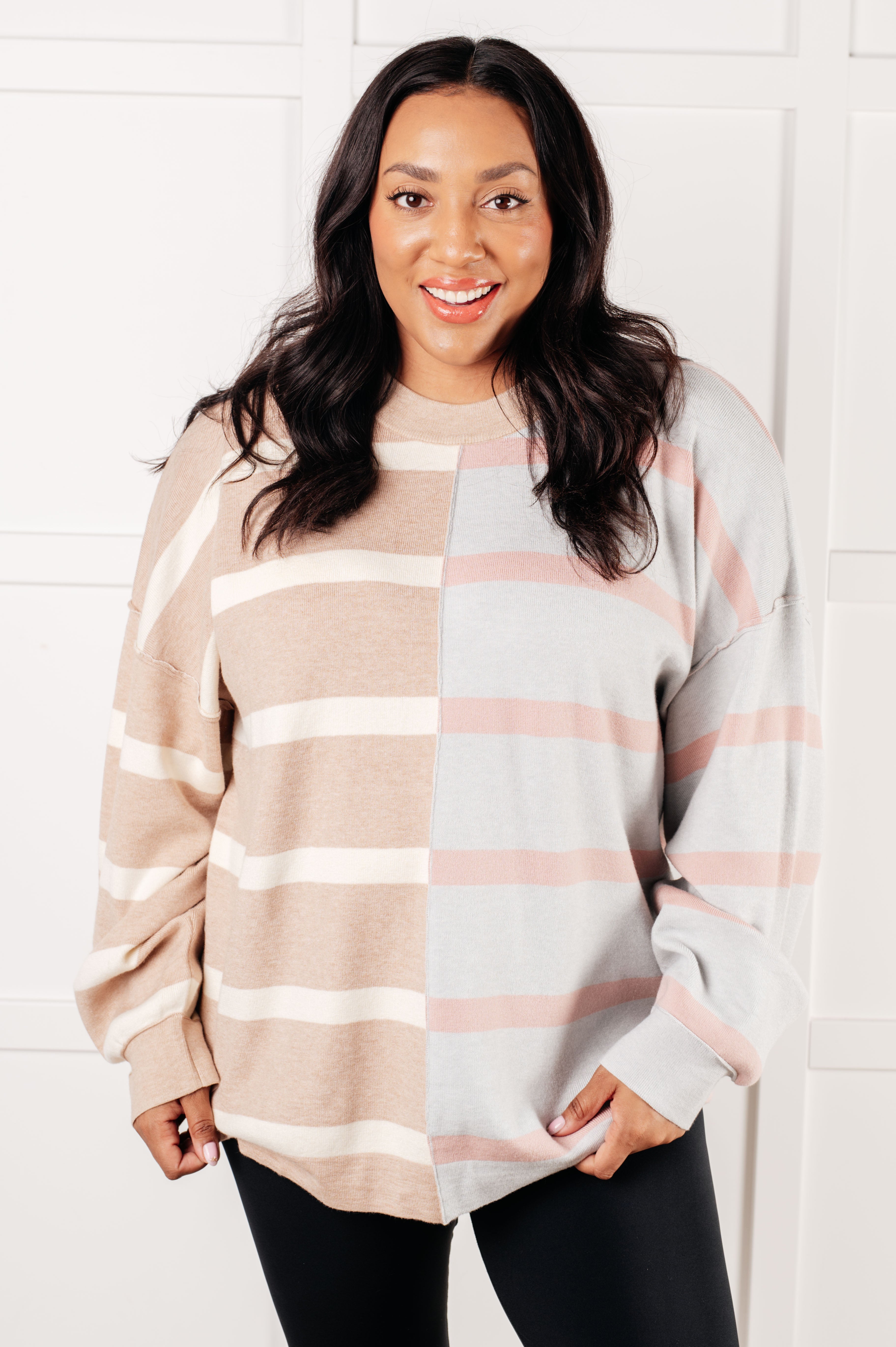 Smiling woman wearing oversized striped patchwork sweater in beige and light gray with pink accents indoors