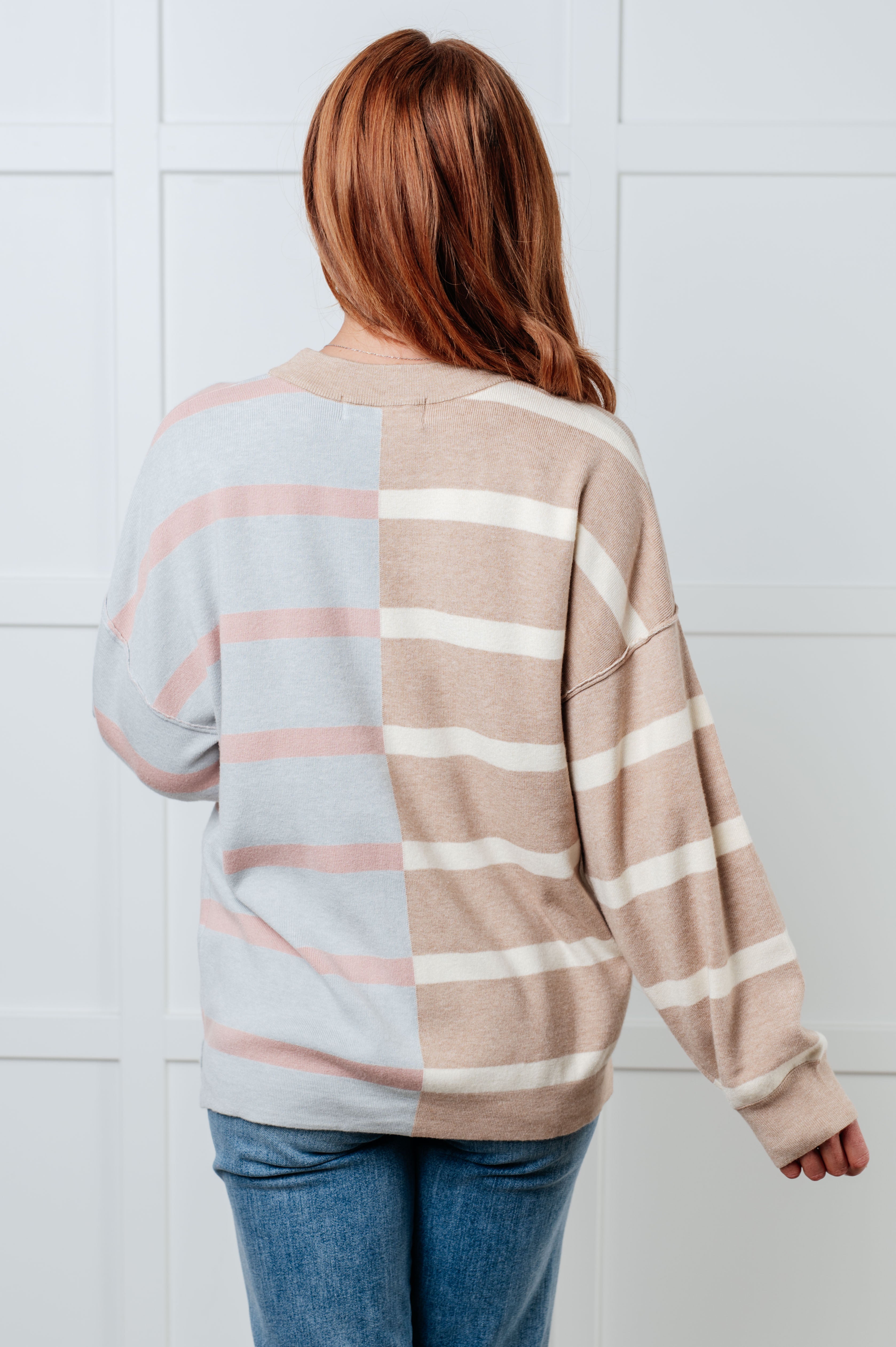 Woman with red hair wearing striped patchwork sweater in beige, cream, and light blue standing indoors against white wall