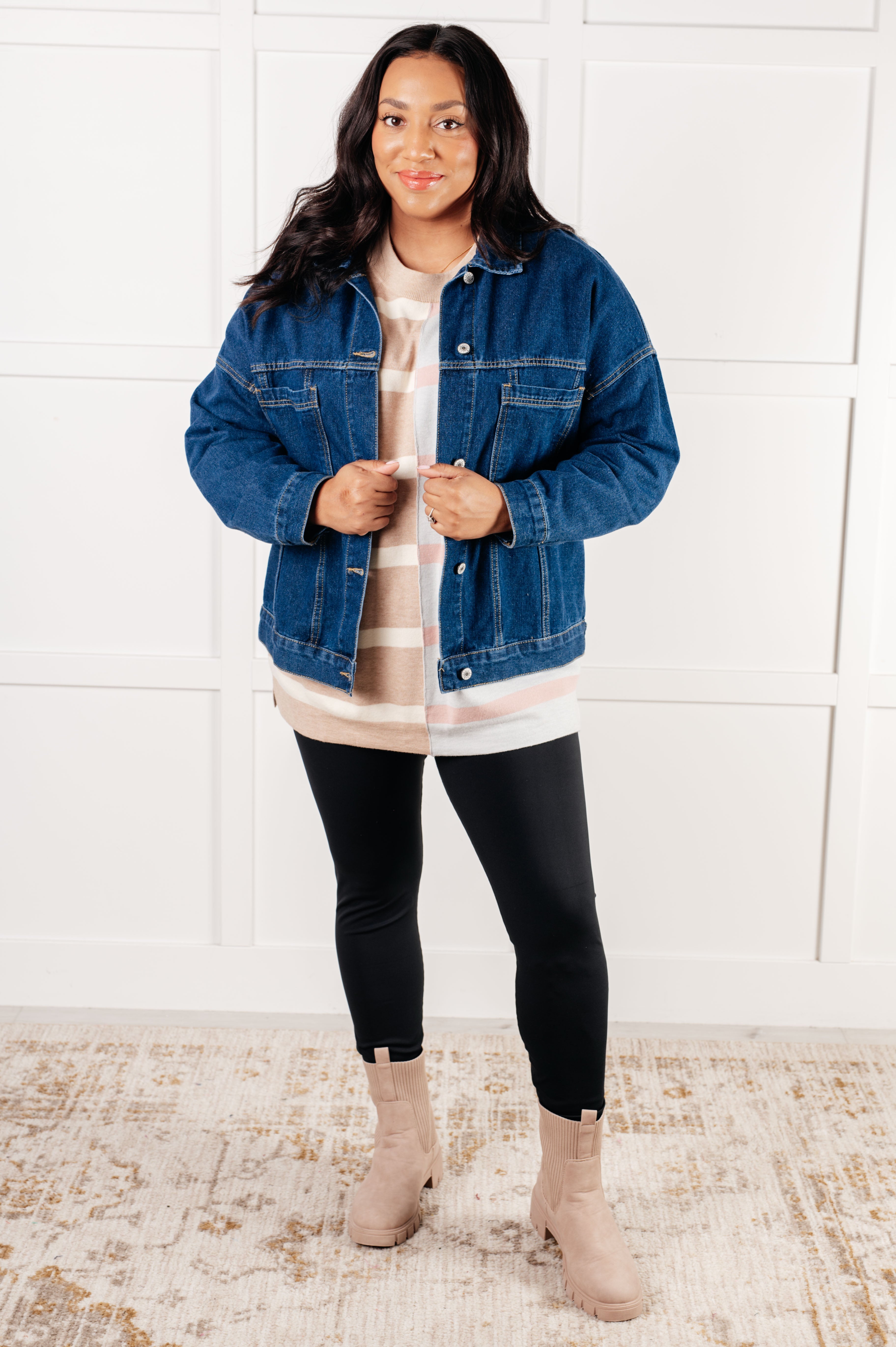 Woman wearing striped patchwork sweater in beige and white under denim jacket with black leggings and beige boots indoors