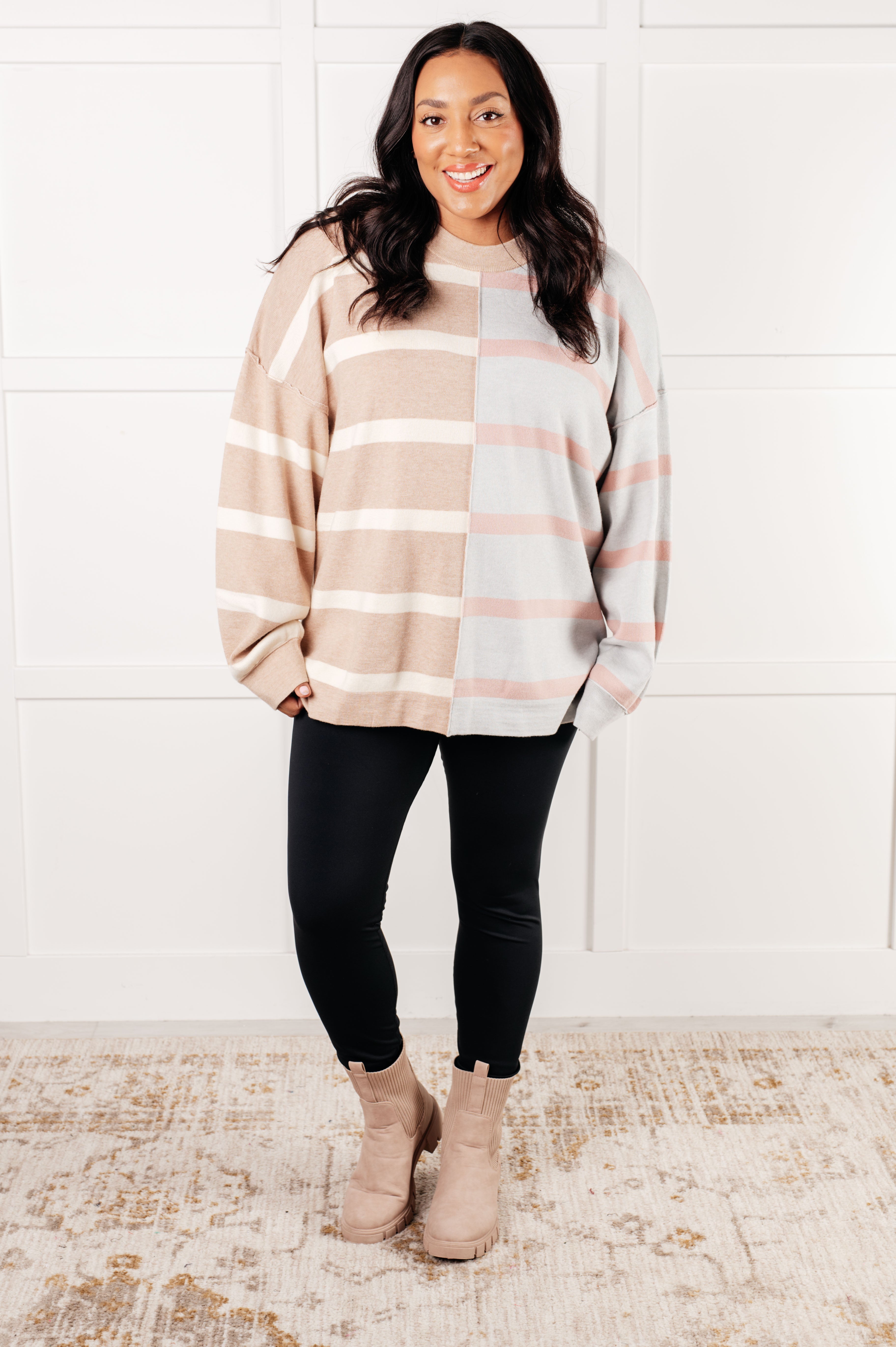 Smiling woman wearing a beige and light gray striped patchwork sweater paired with black leggings and beige boots indoors