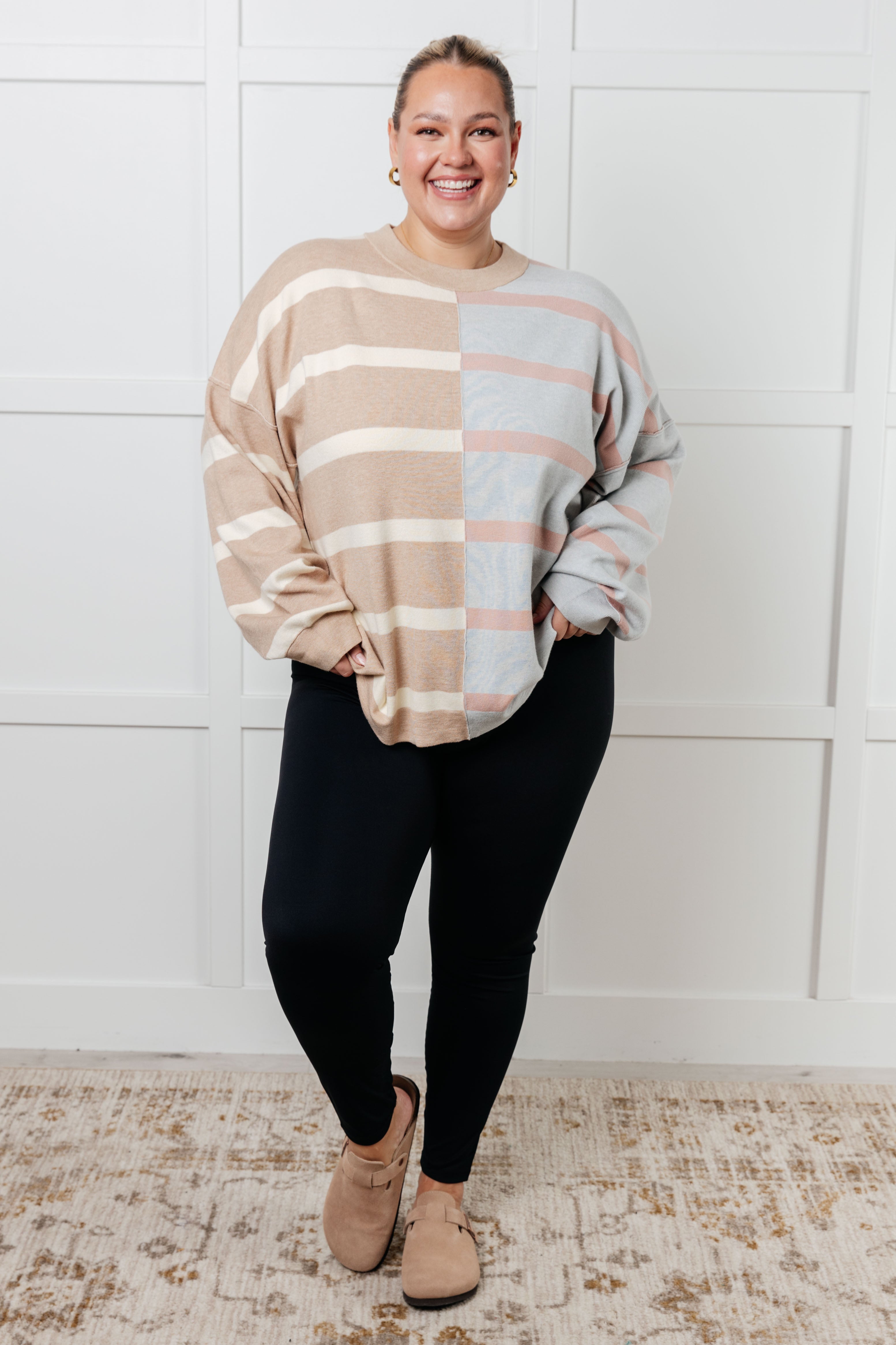 Plus-size woman smiling wearing oversized striped patchwork sweater in beige, light blue, and pink with black leggings indoors