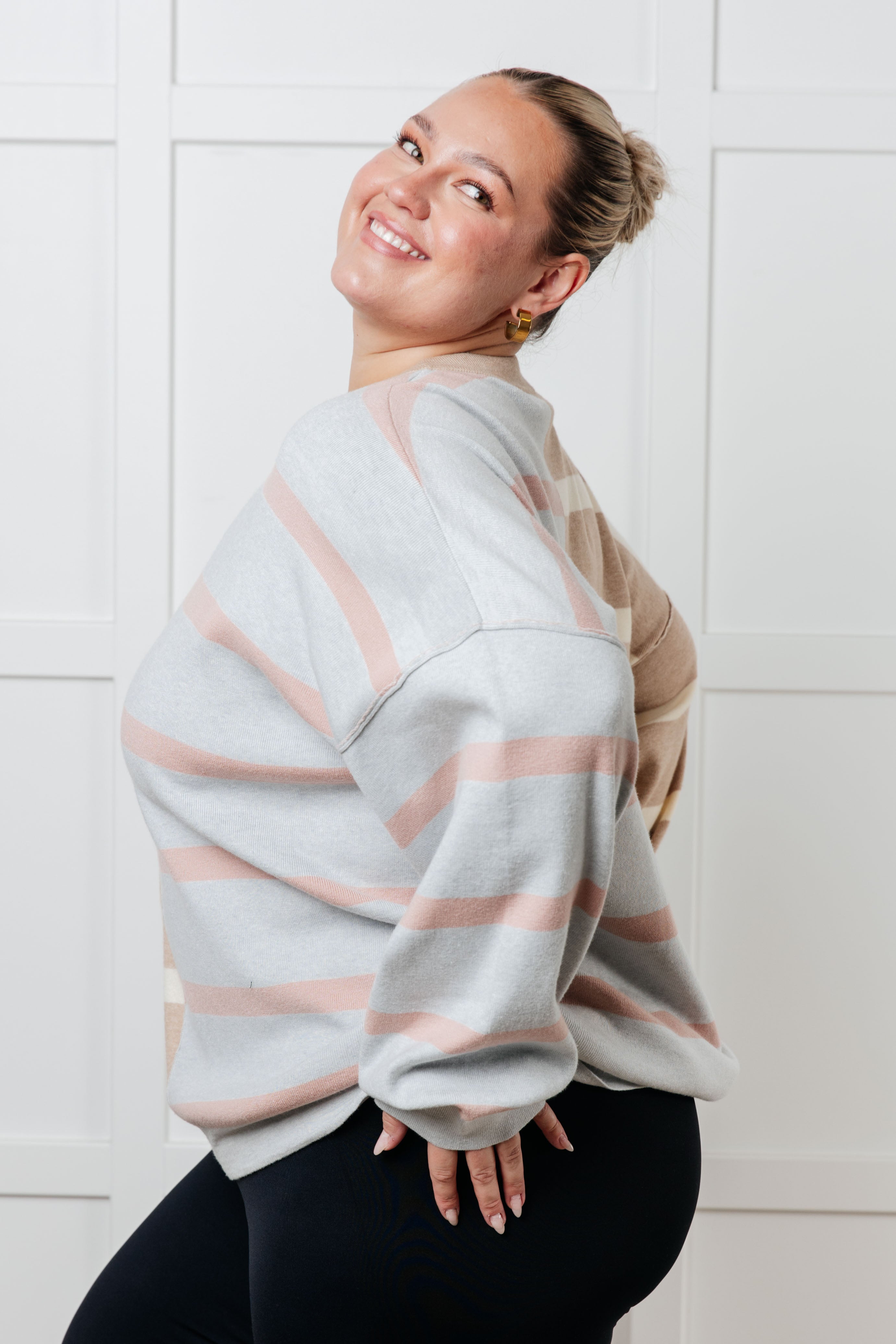 Smiling woman wearing oversized striped patchwork sweater in soft pink and gray posing indoors against white paneled wall