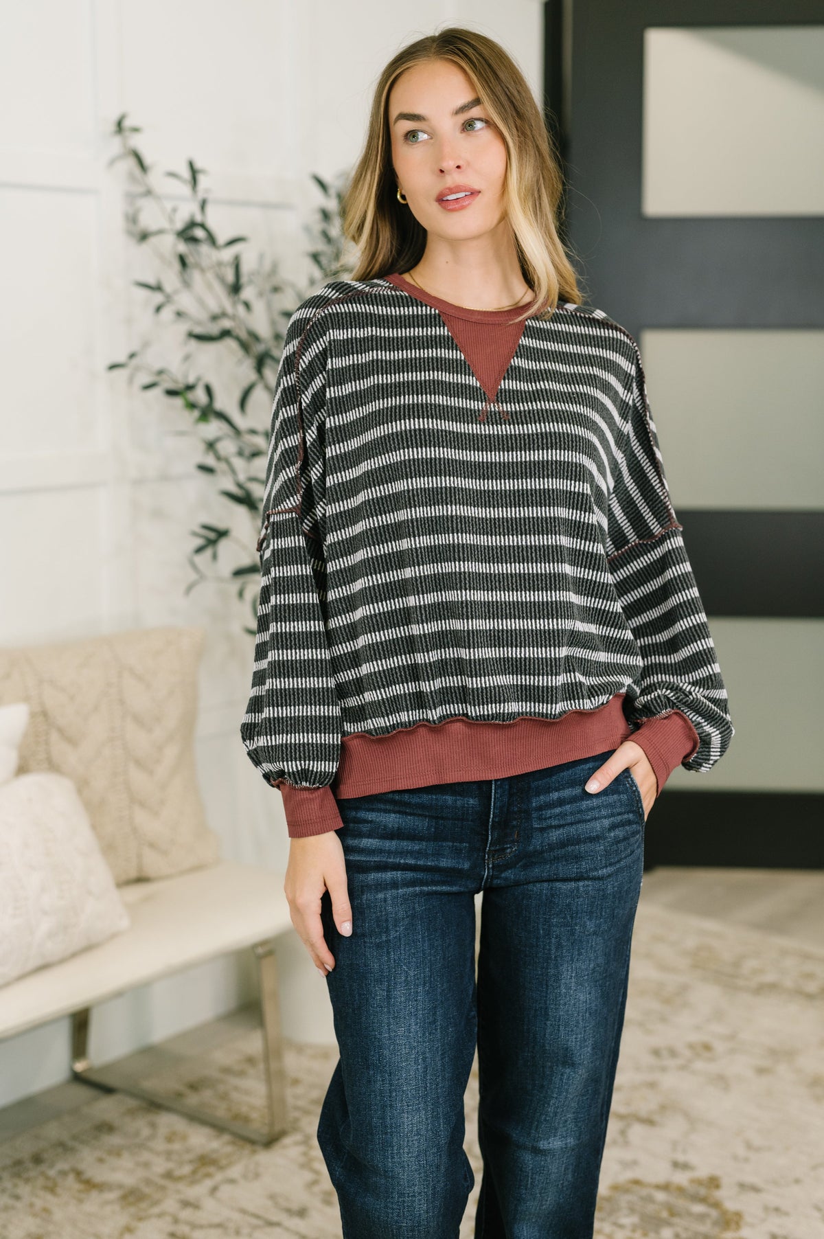 Woman wearing a charcoal contrast textured knit pullover over a rust top with dark jeans in a bright home interior