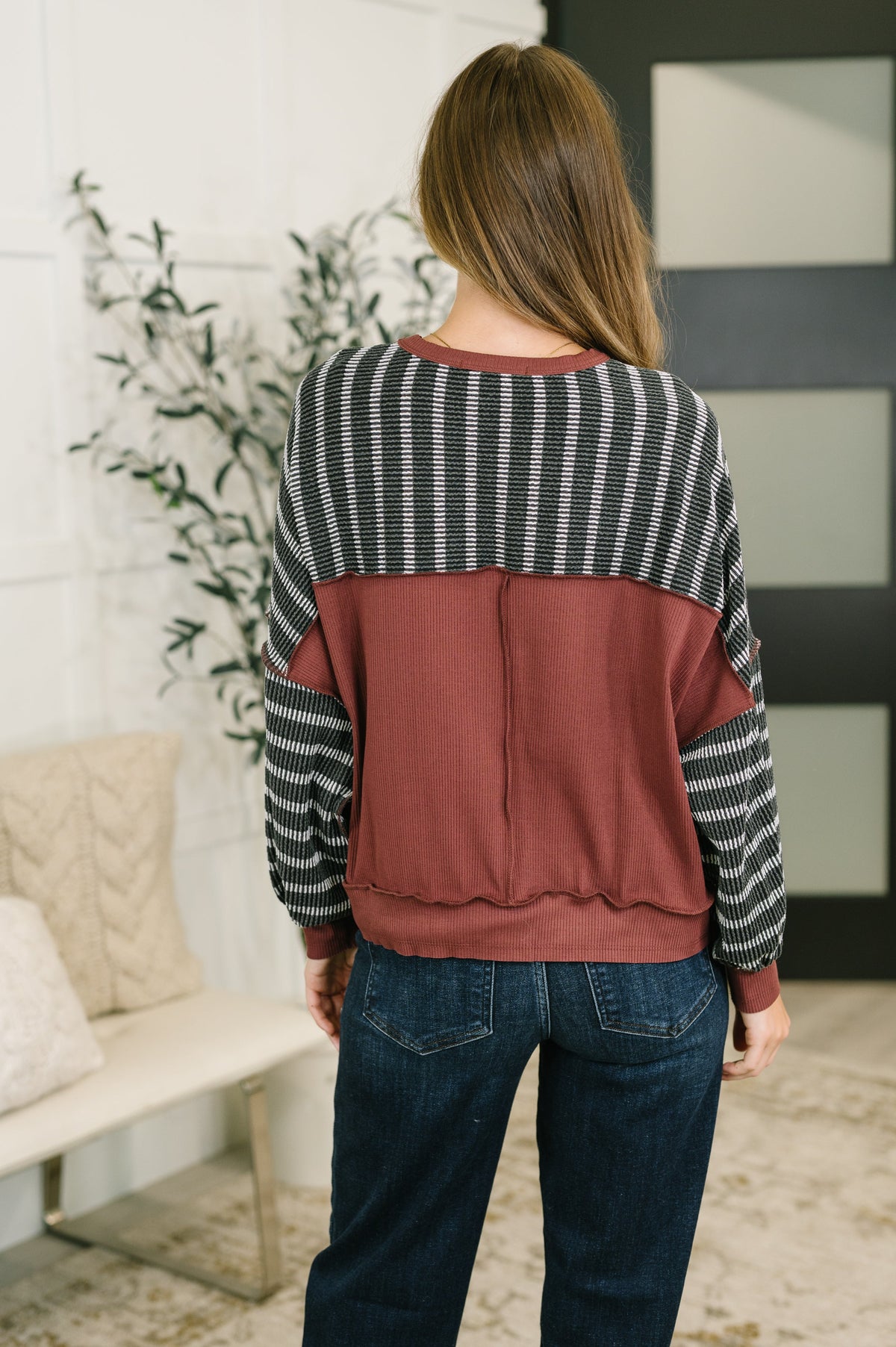 Woman viewed from behind wearing a charcoal contrast textured knit pullover with a rust panel and blue jeans in a cozy home interior