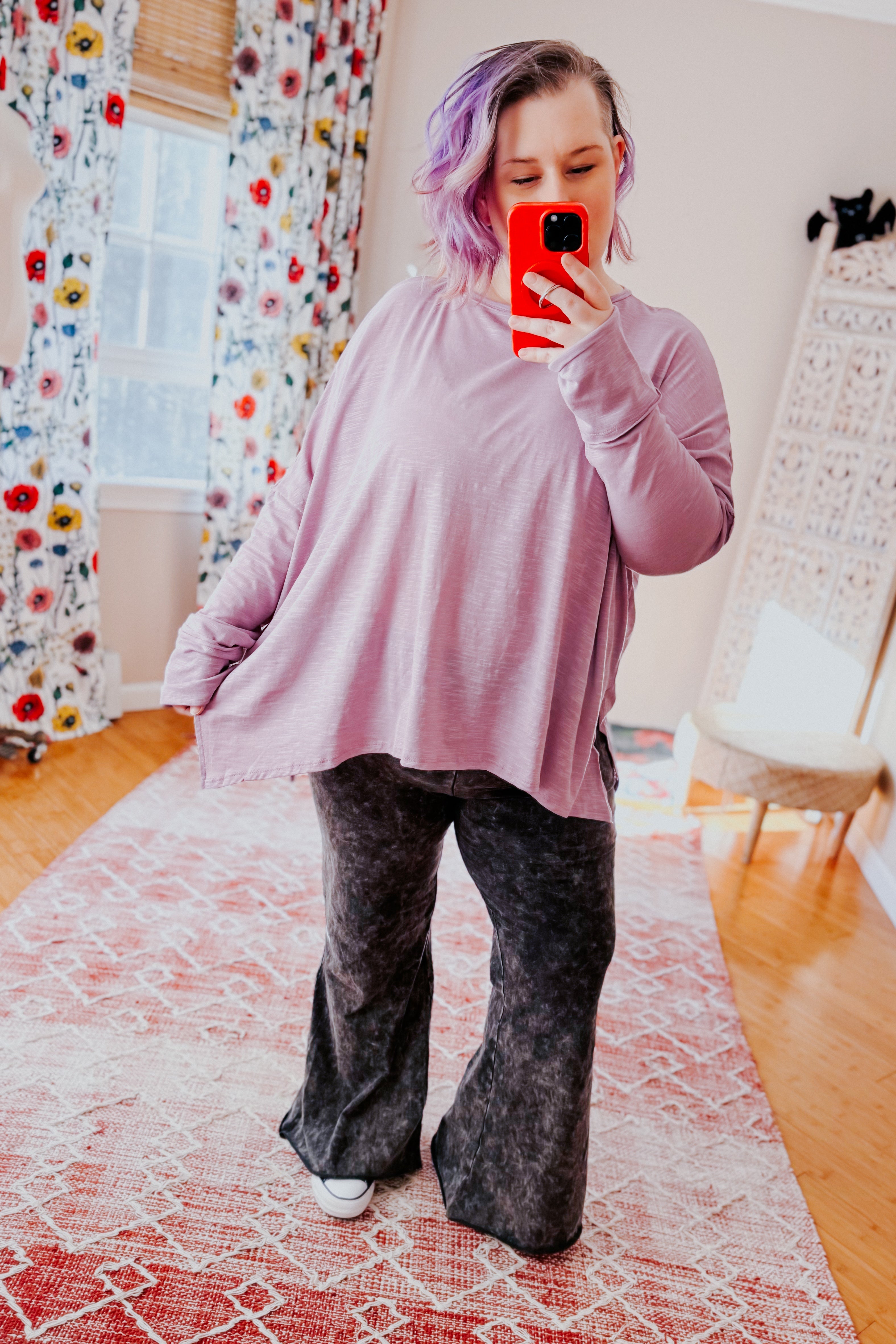 Woman wearing a lavender sweater and gray mineral wash pants taking a mirror selfie in a room with floral curtains.