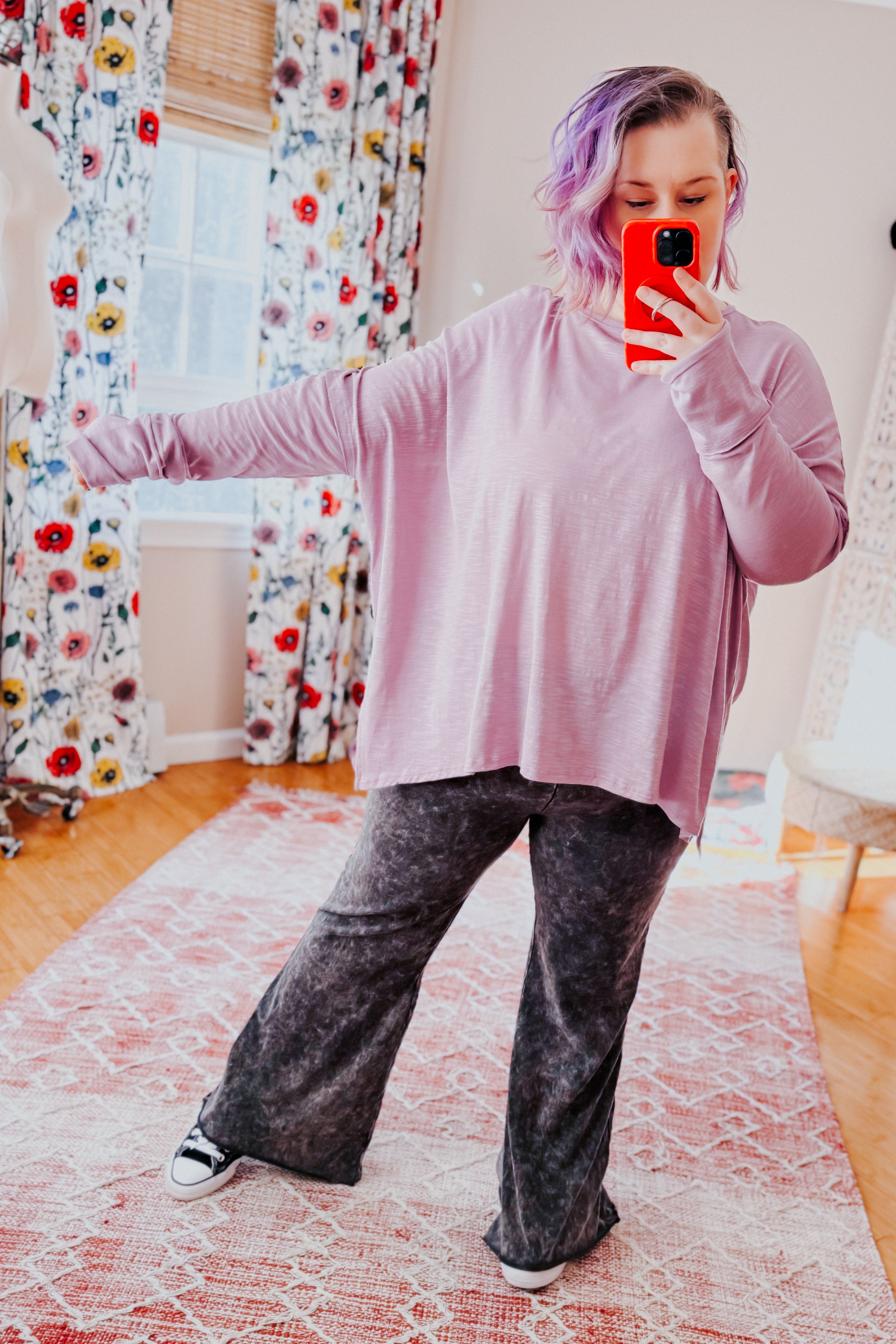 Woman wearing a lavender sweater and gray mineral wash pants taking a mirror selfie in a room with floral curtains.