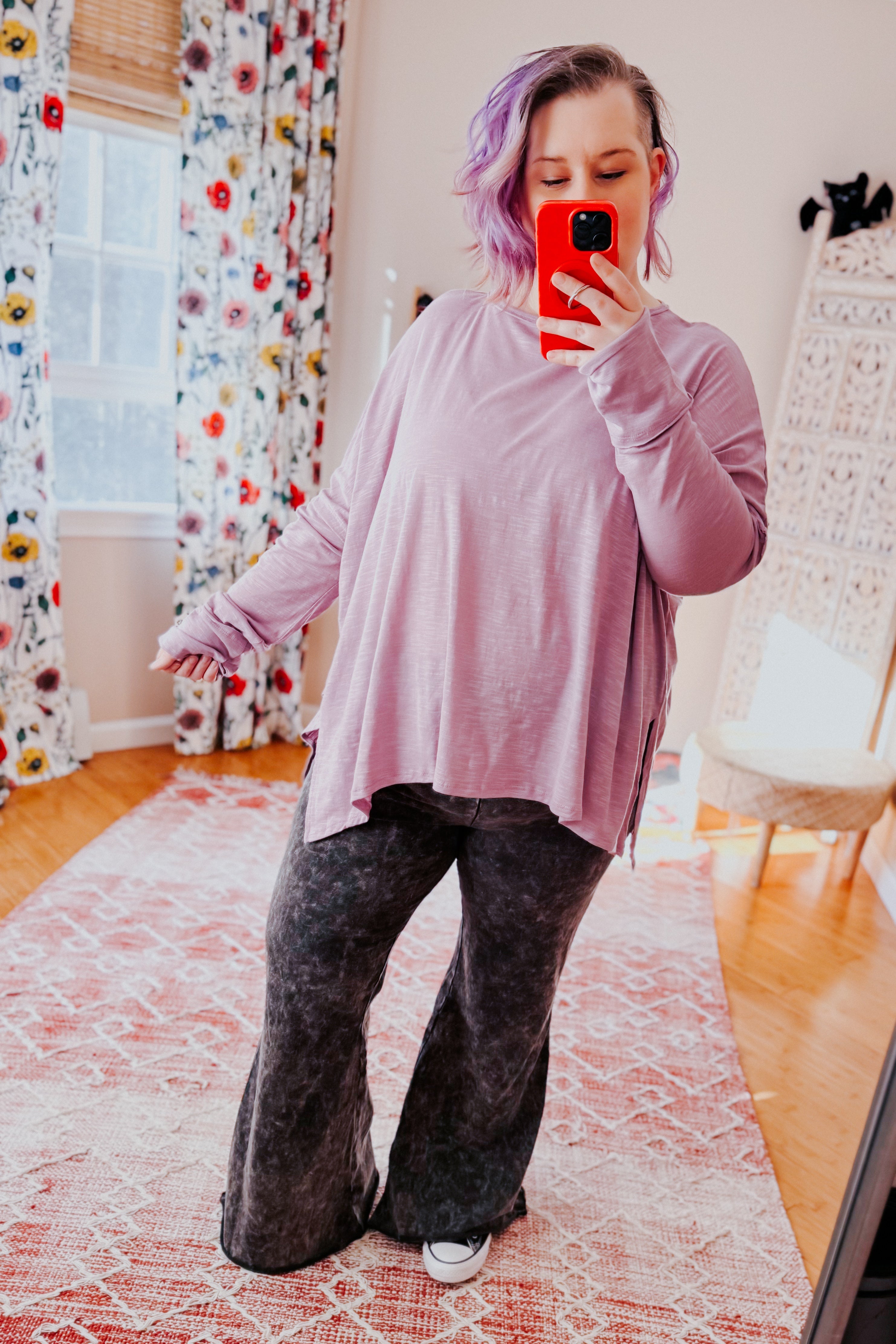 Woman wearing a lavender sweater and gray mineral wash pants taking a mirror selfie in a room with floral curtains.