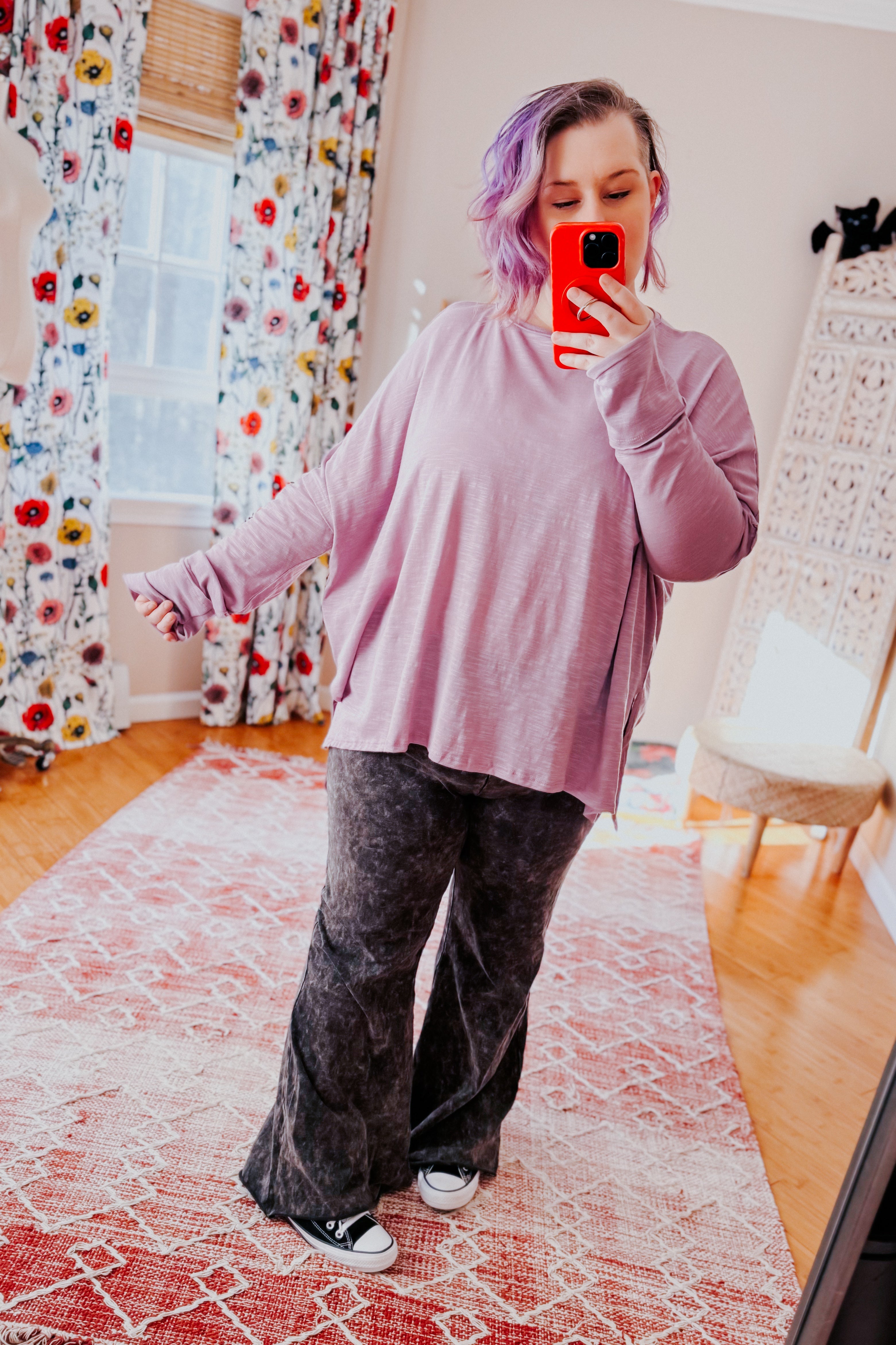 Woman wearing a lavender sweater and gray mineral wash pants taking a mirror selfie in a room with floral curtains.