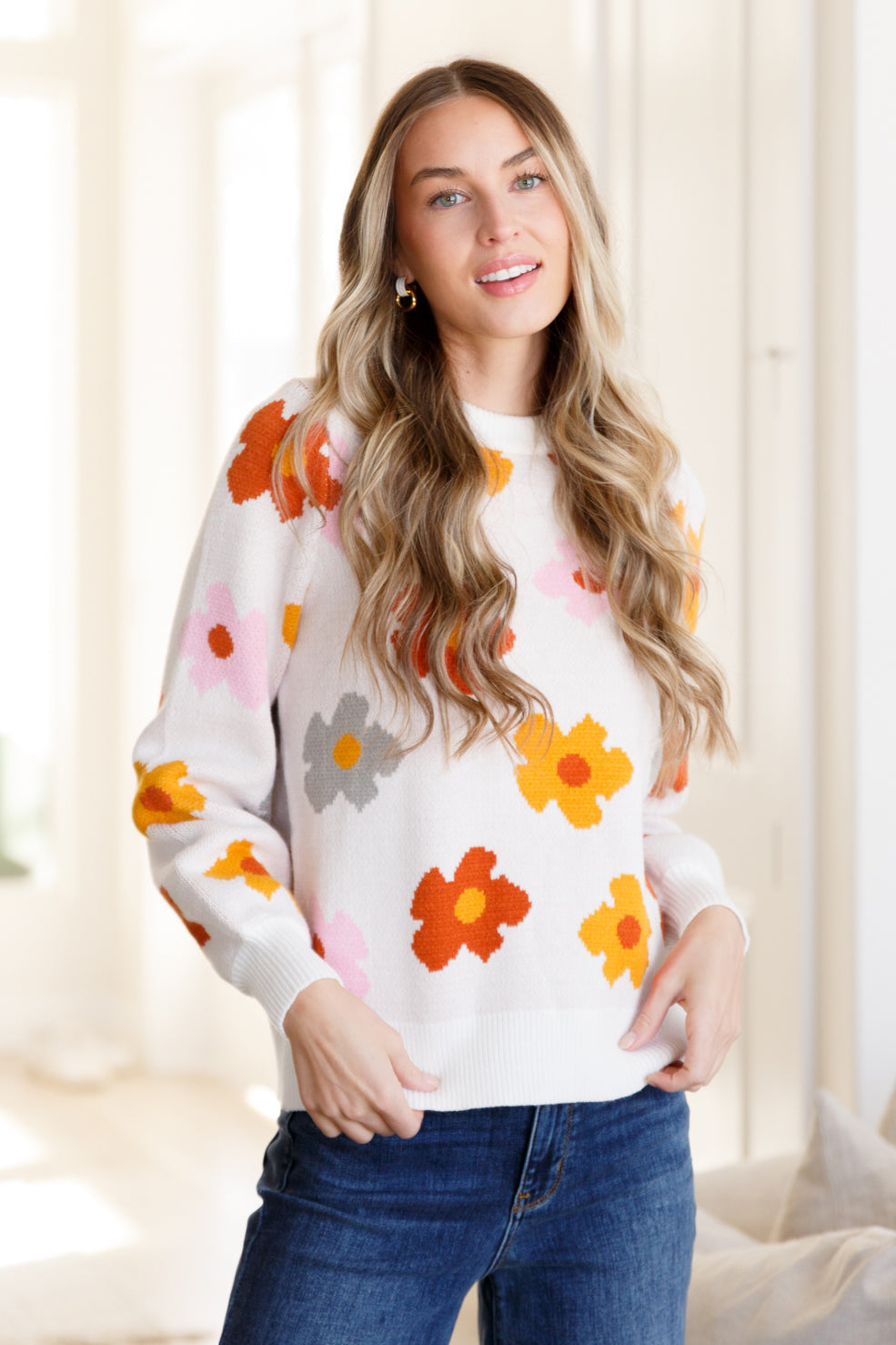 Woman wearing white sweater with bright orange, yellow, pink, and gray floral patterns paired with blue jeans indoors