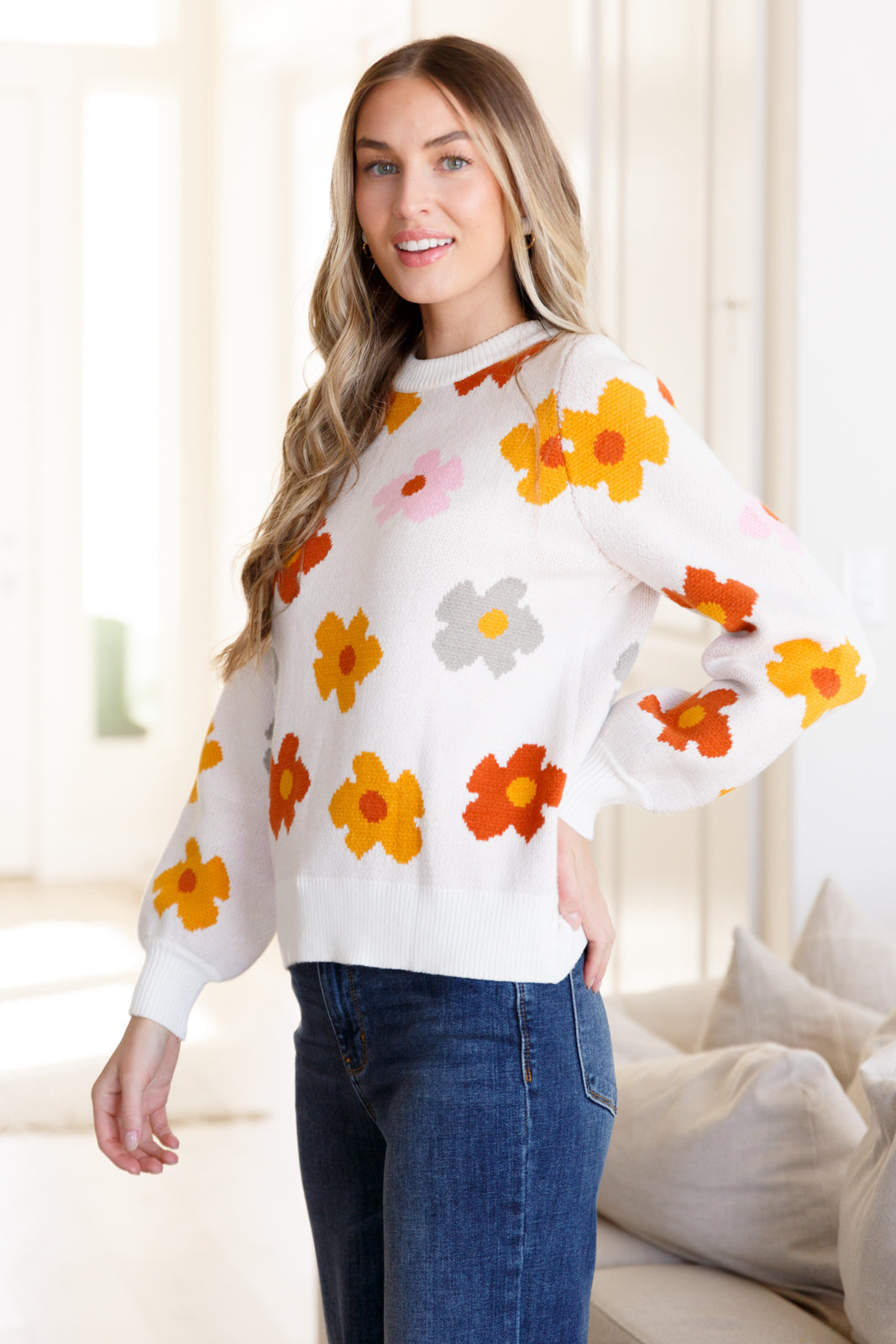 Woman wearing cream sweater with orange, yellow, pink, and gray floral patterns paired with dark blue jeans indoors