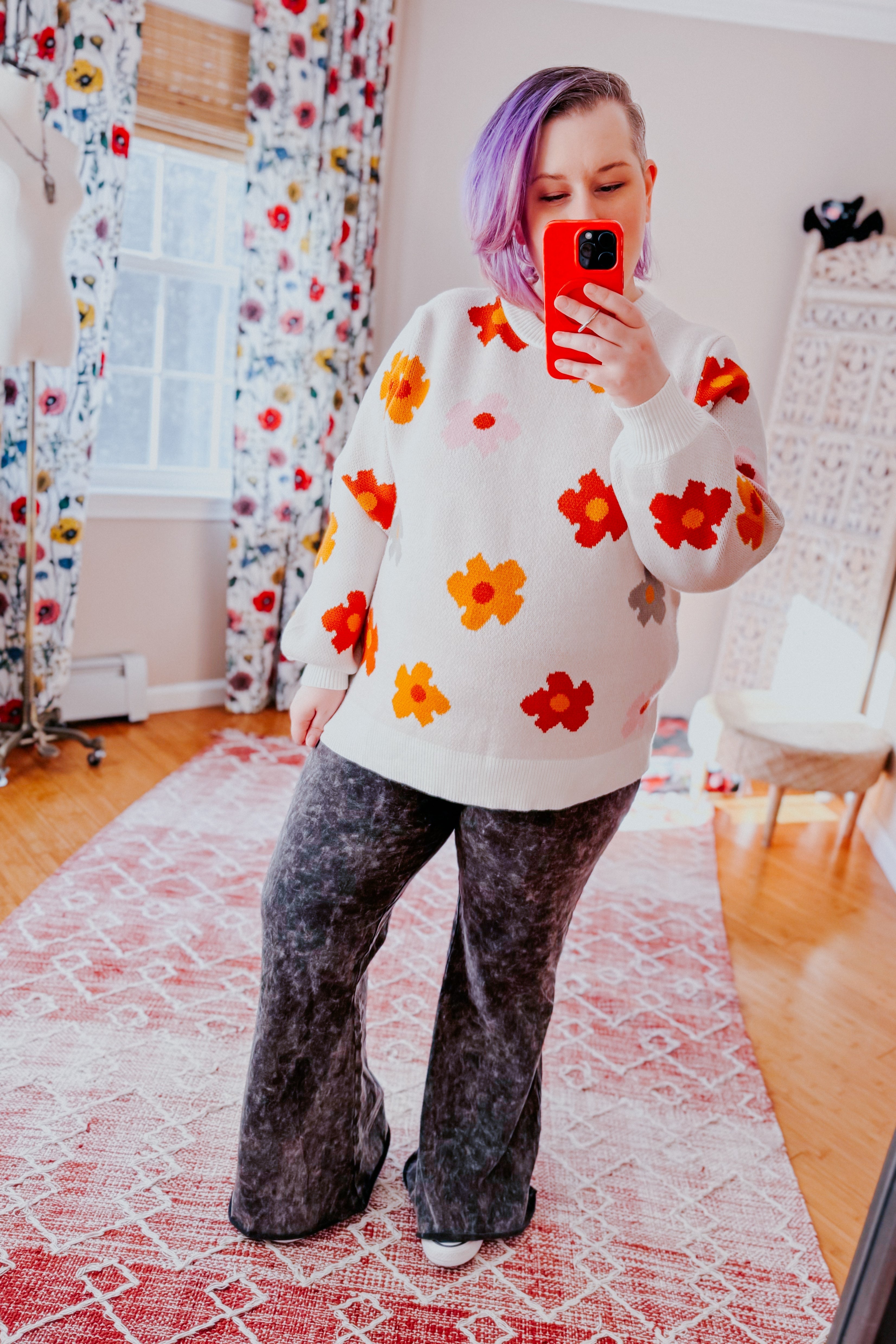 Woman with purple hair wearing white sweater with bold orange and red flowers paired with black patterned wide-leg pants indoors