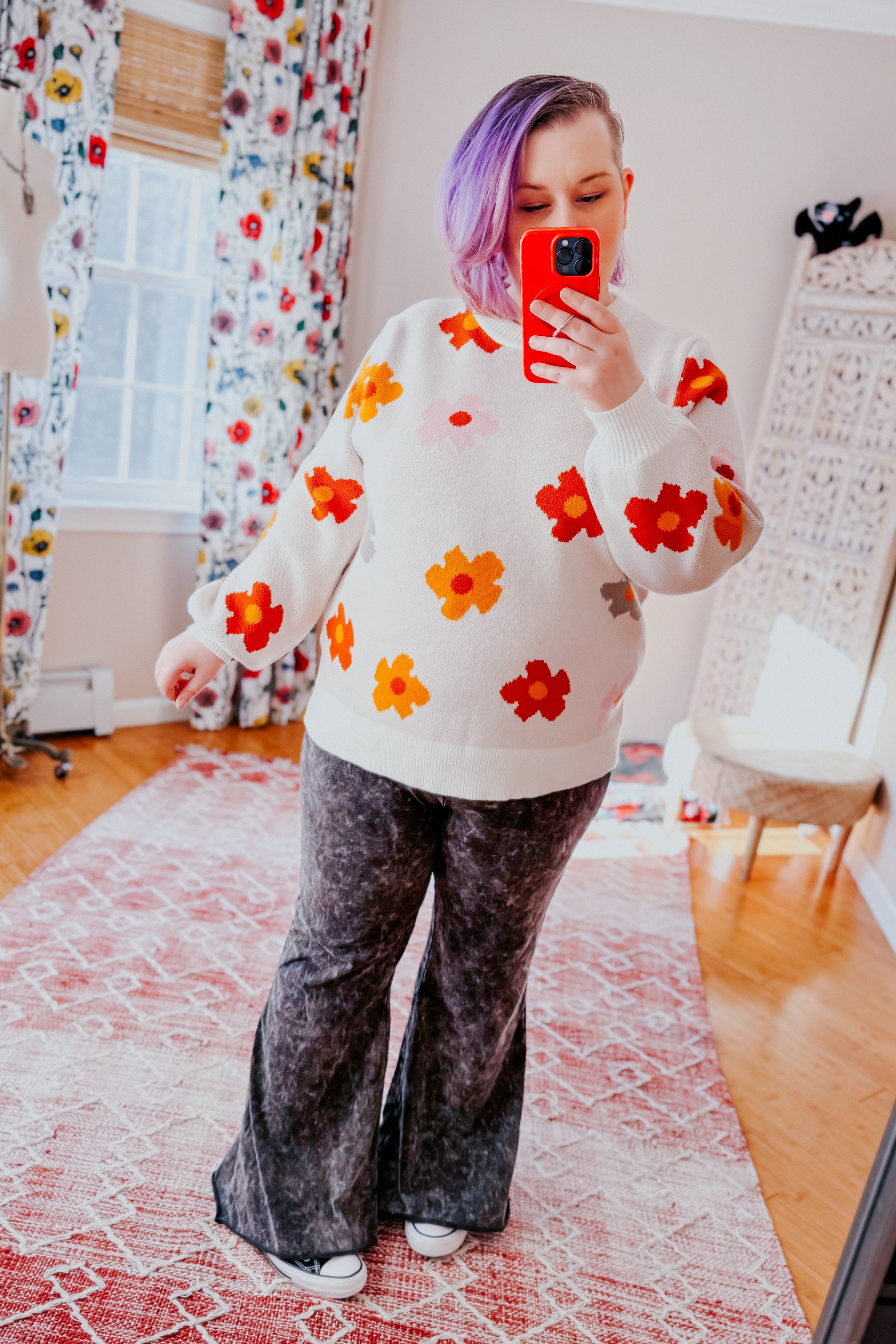 Woman with purple hair wearing white sweater with red and orange floral print and dark gray wide-leg pants indoors