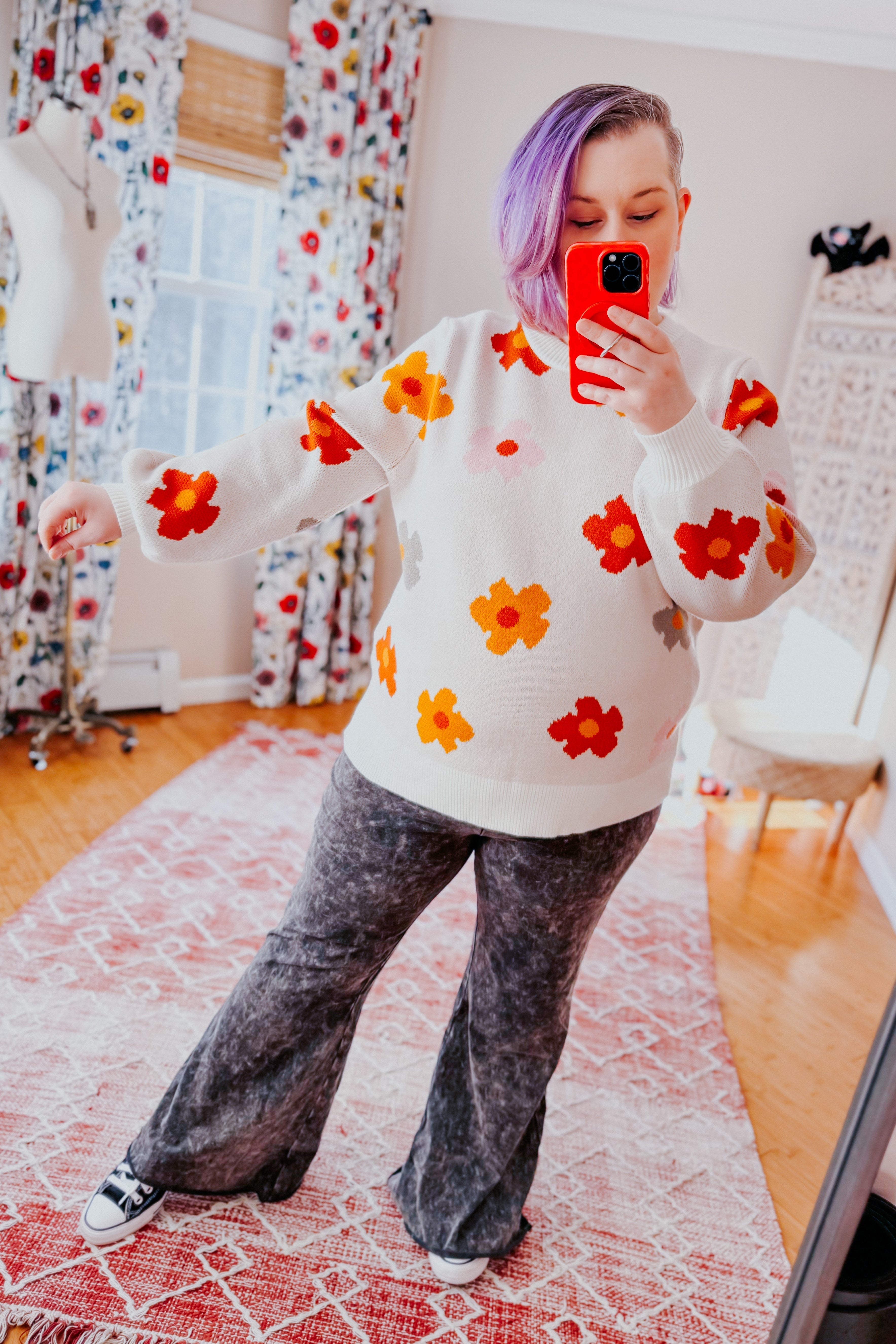 Woman with short purple hair wearing white sweater with large red and orange flowers and gray flare pants indoors