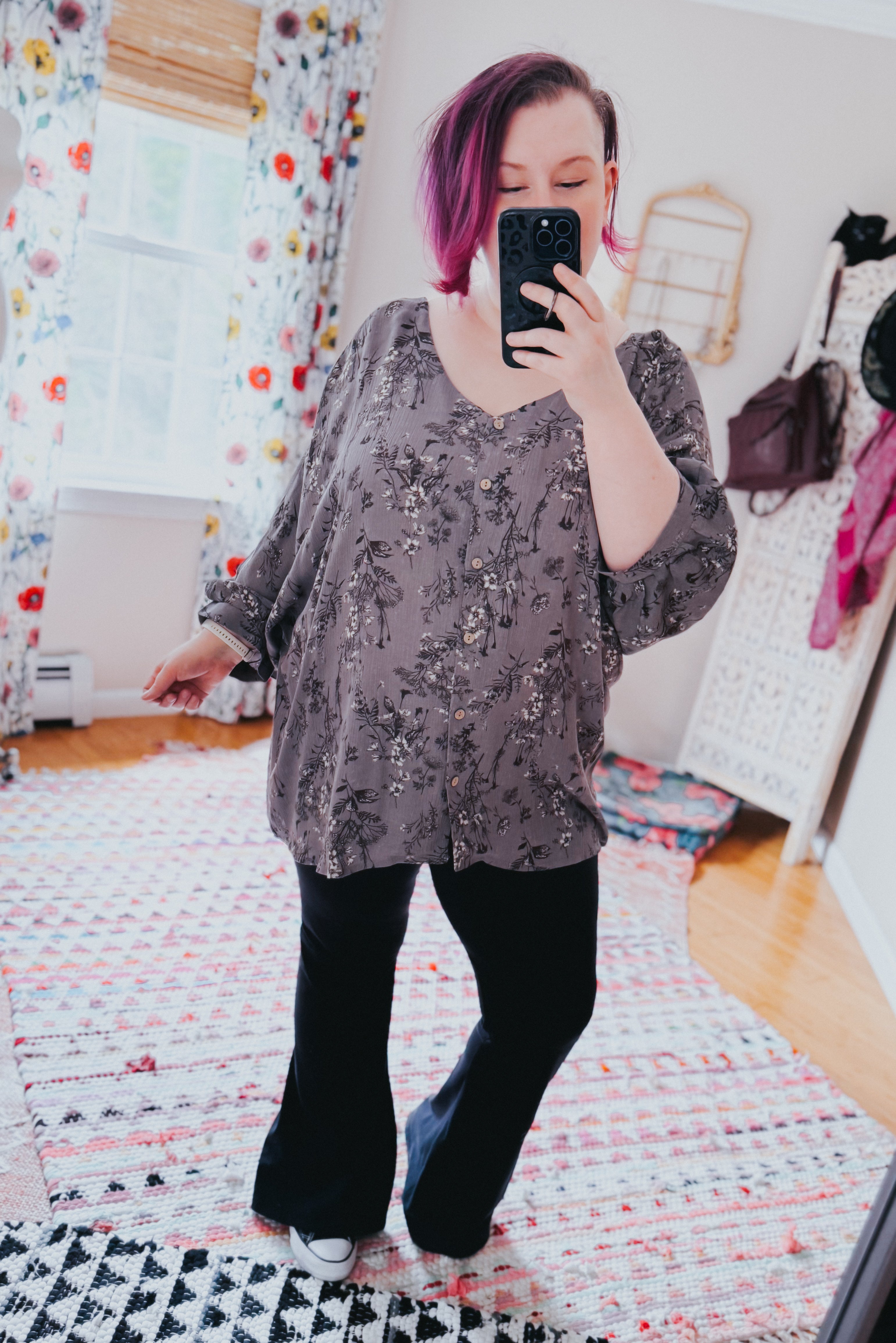 Woman with short purple hair wearing loose dove grey floral crinkle blouse paired with black pants taking mirror selfie indoors
