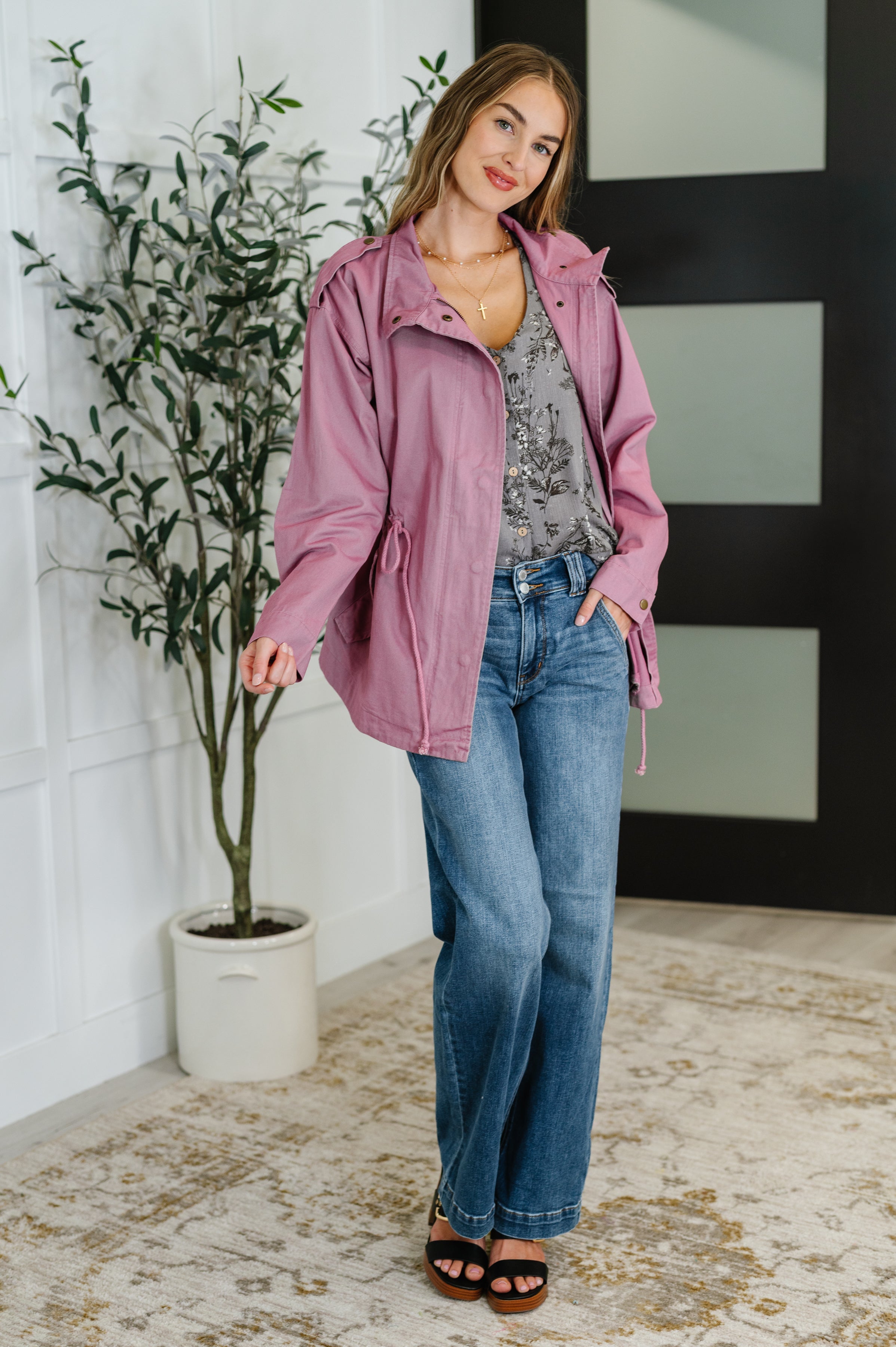 Woman wearing muted mauve jacket over grey printed blouse with blue jeans and black sandals indoors by a potted plant