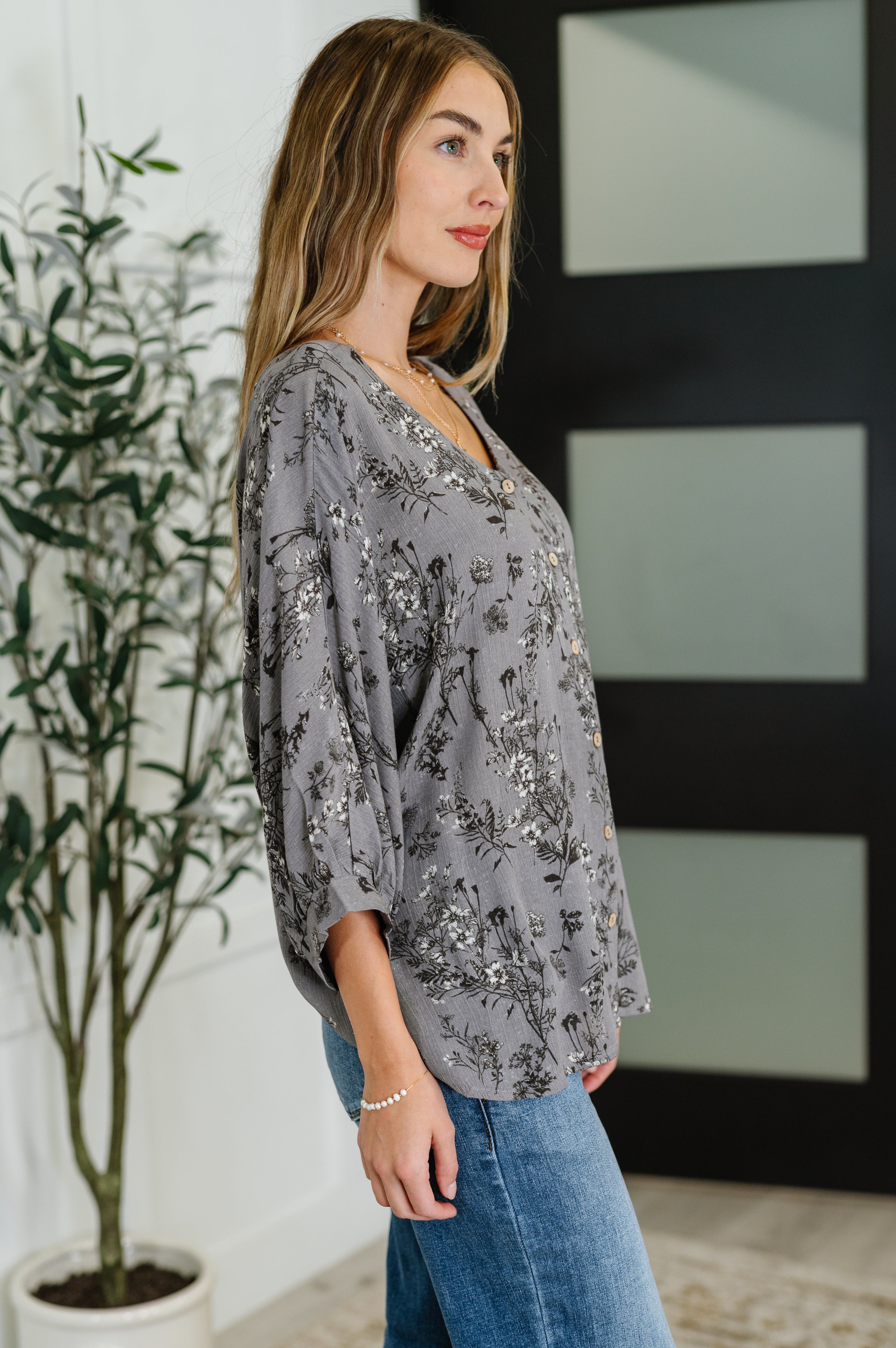 Woman wearing loose dune grey crinkle woven blouse with black and white floral print and blue jeans indoors
