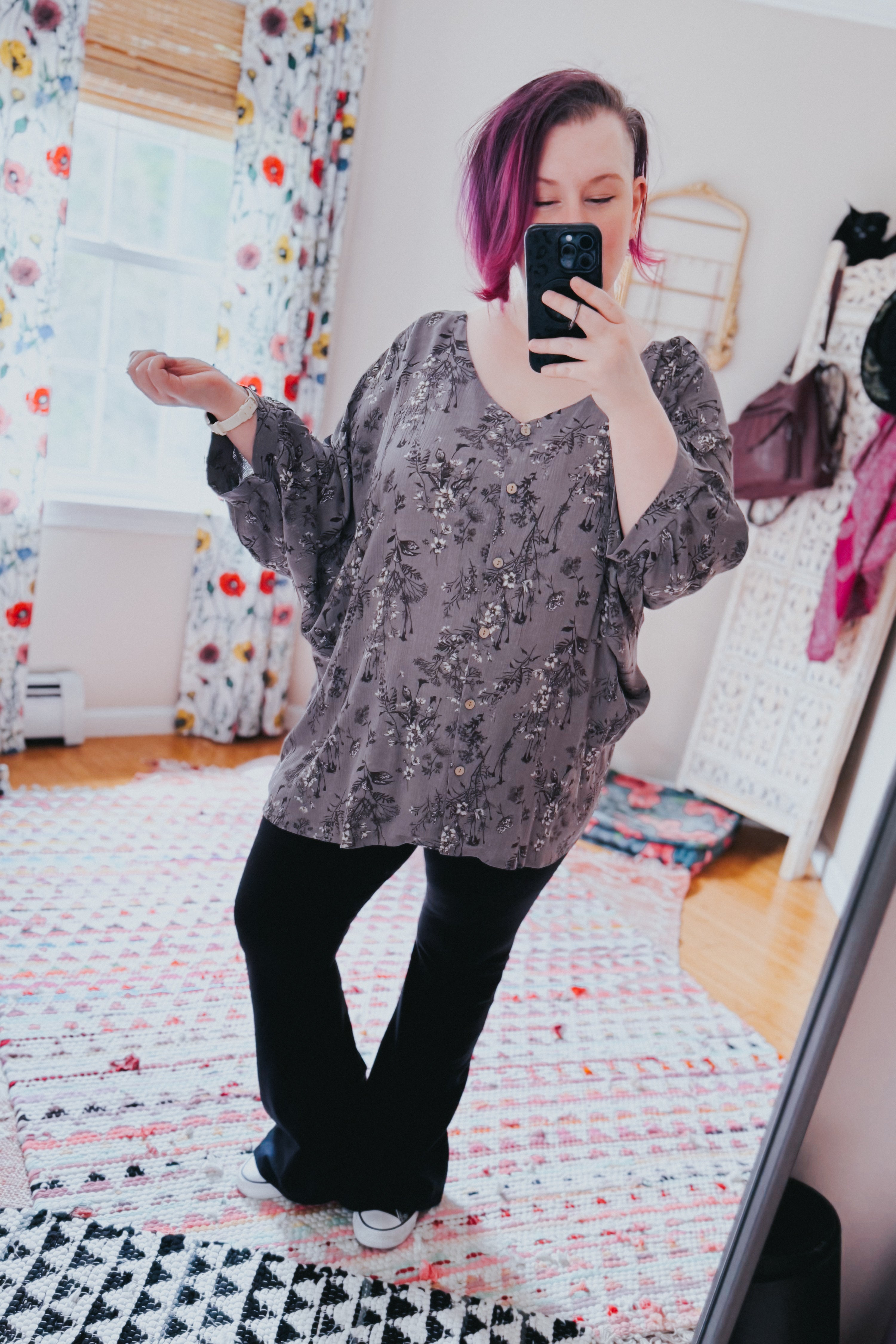 Woman with short purple hair wearing loose dune grey crinkle blouse with subtle floral print and black pants taking mirror selfie indoors
