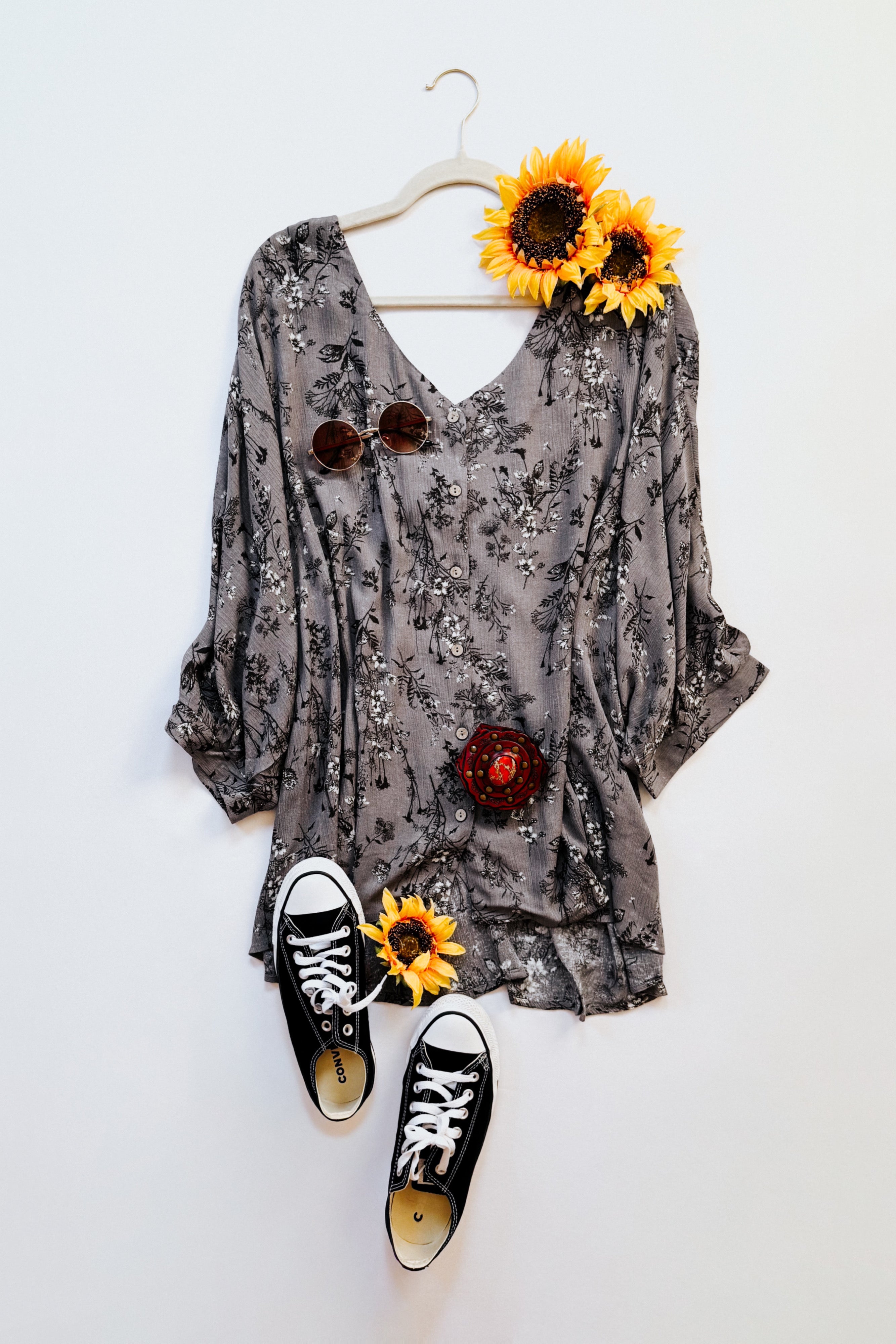 Dune grey crinkled blouse with black floral print styled with brown sunglasses, black sneakers, and sunflowers on white background