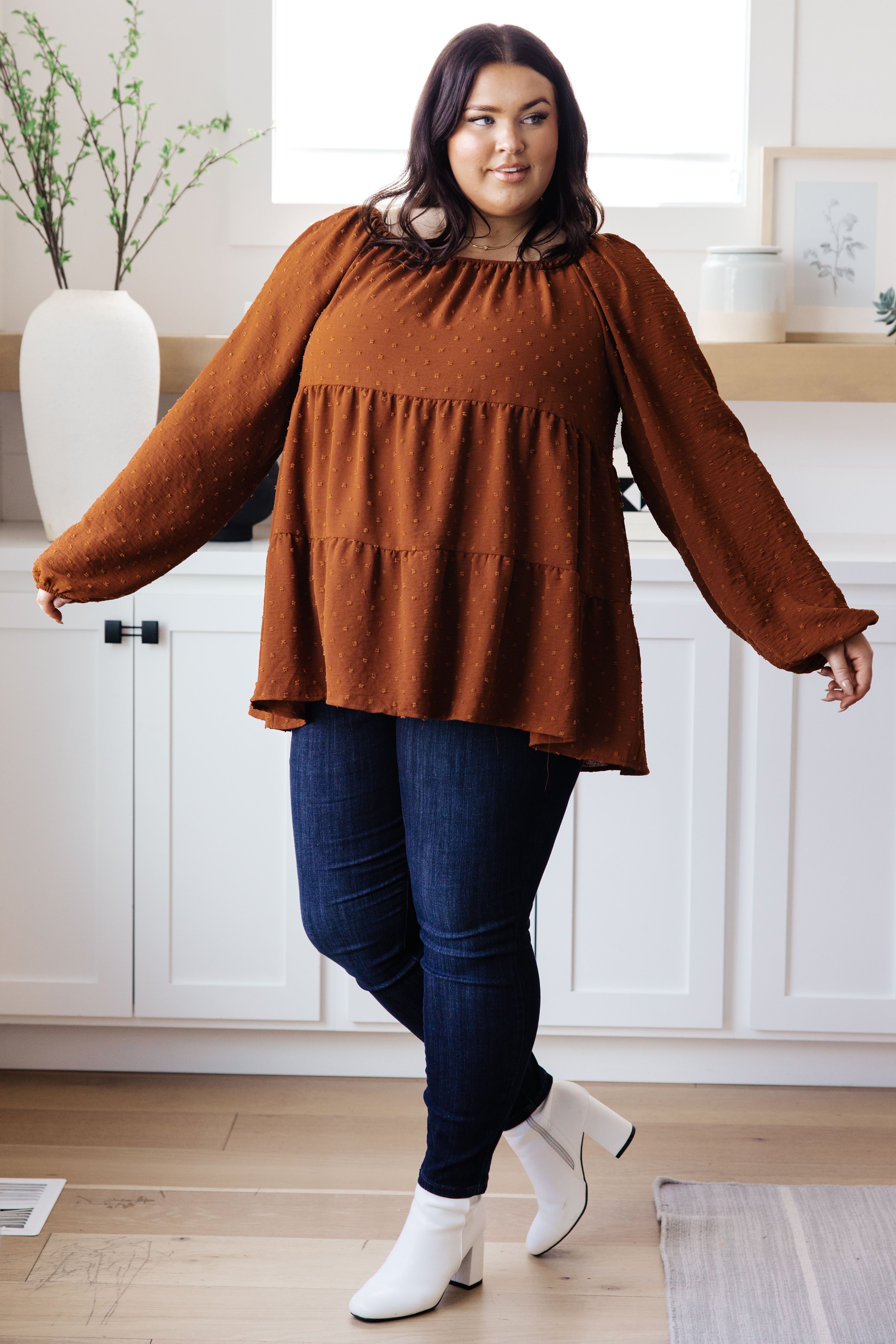 Plus-size model wearing rust tiered babydoll top with puffed sleeves paired with dark skinny jeans and white ankle boots indoors