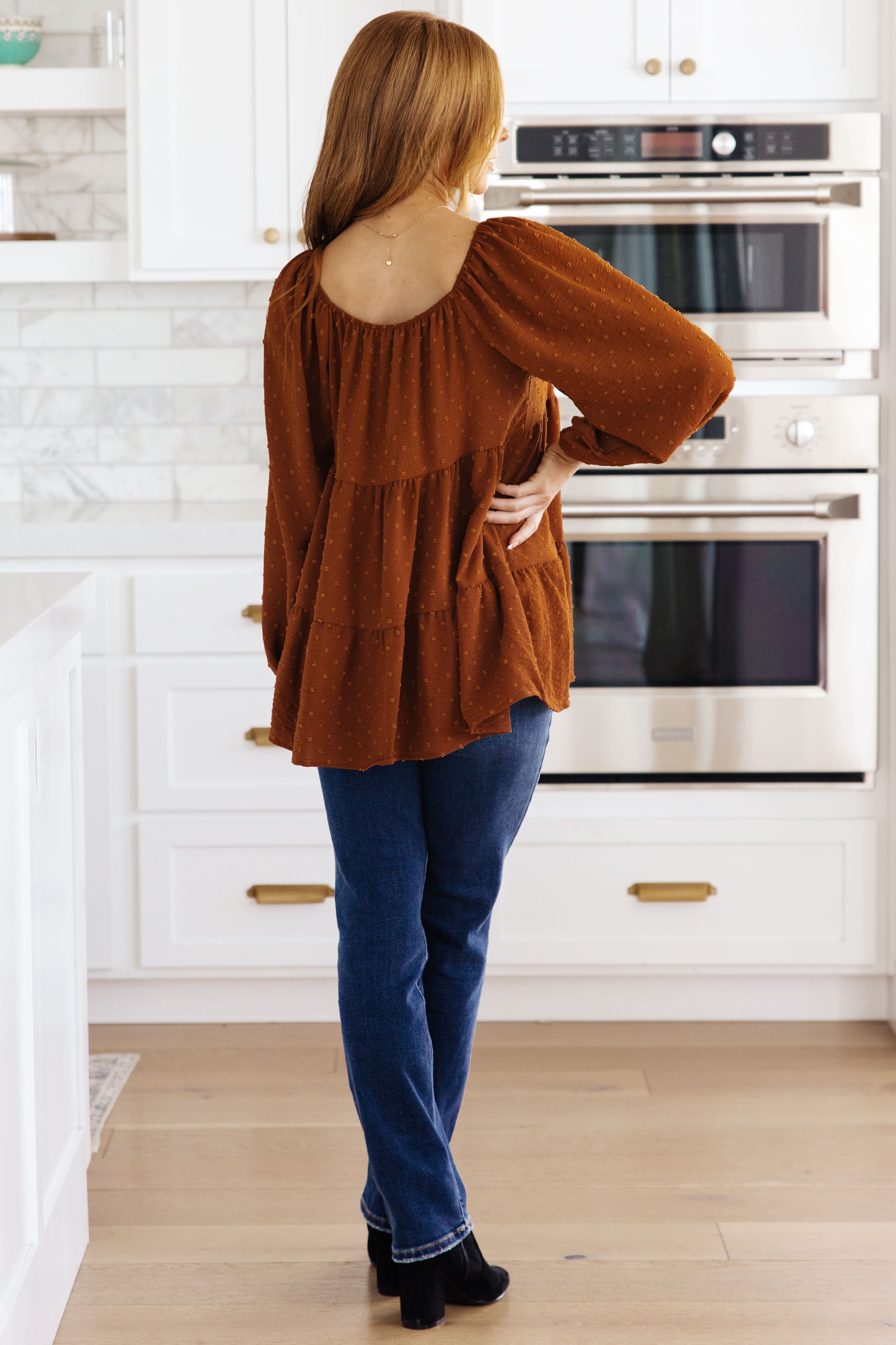 Woman wearing rust textured tiered babydoll top with long sleeves paired with dark blue jeans standing indoors
