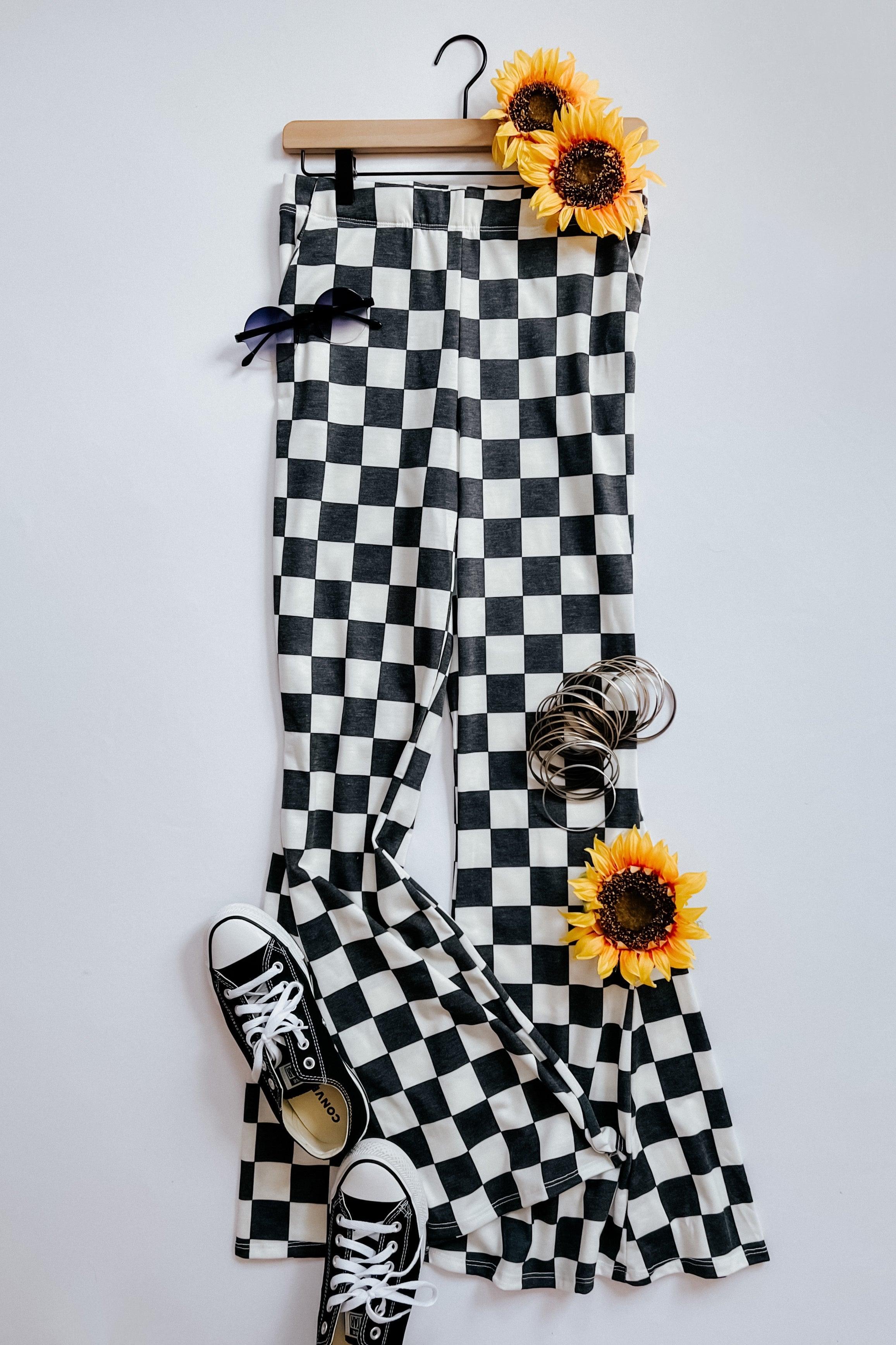 Black and white checkered flared pants on hanger with sneakers, sunglasses, bangles, and sunflowers on white background